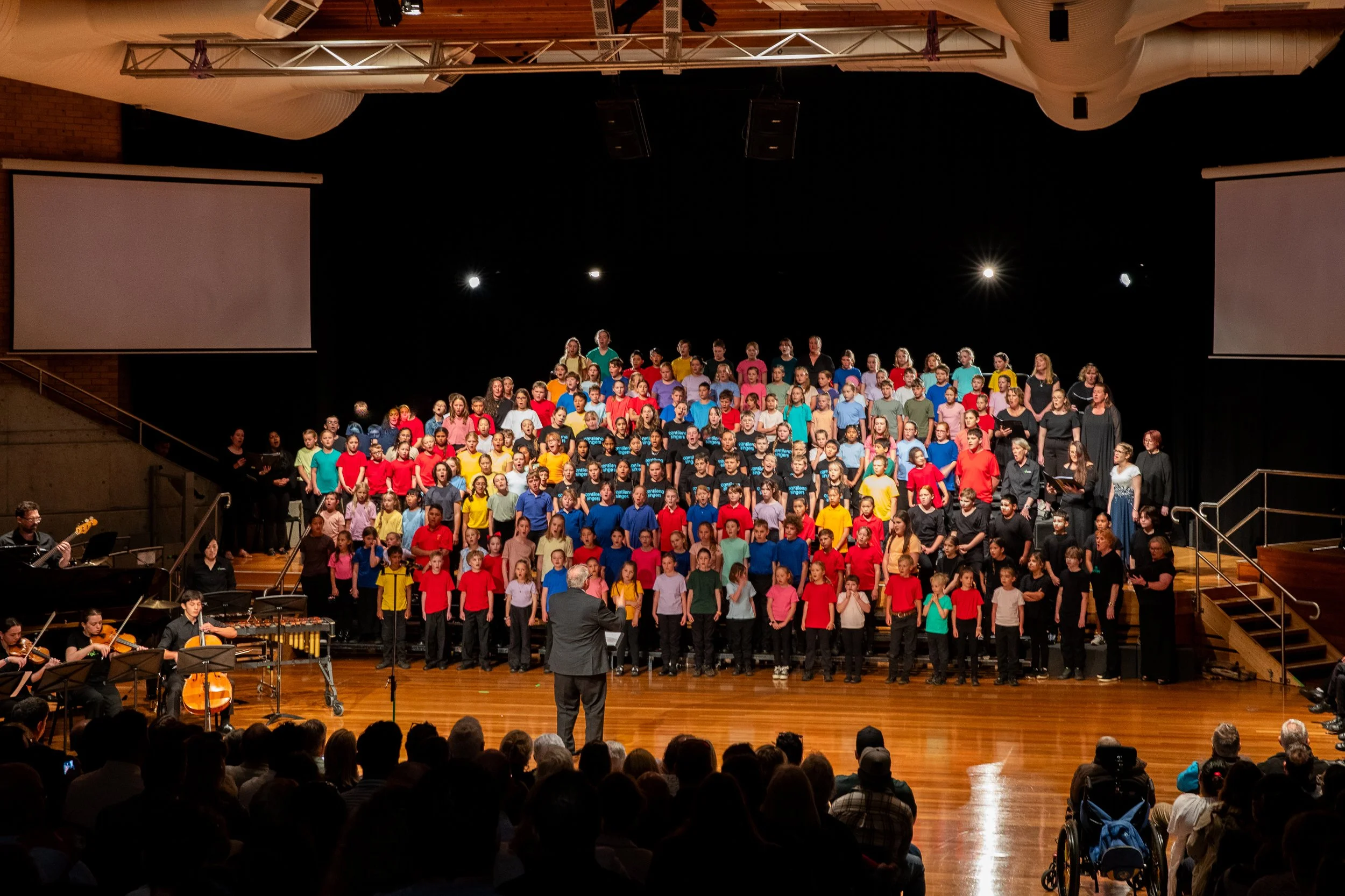 A large choir of children and adults on stage during a performance, directed by a conductor in front of them, with musicians playing instruments on the left, and an audience watching in a concert hall.
