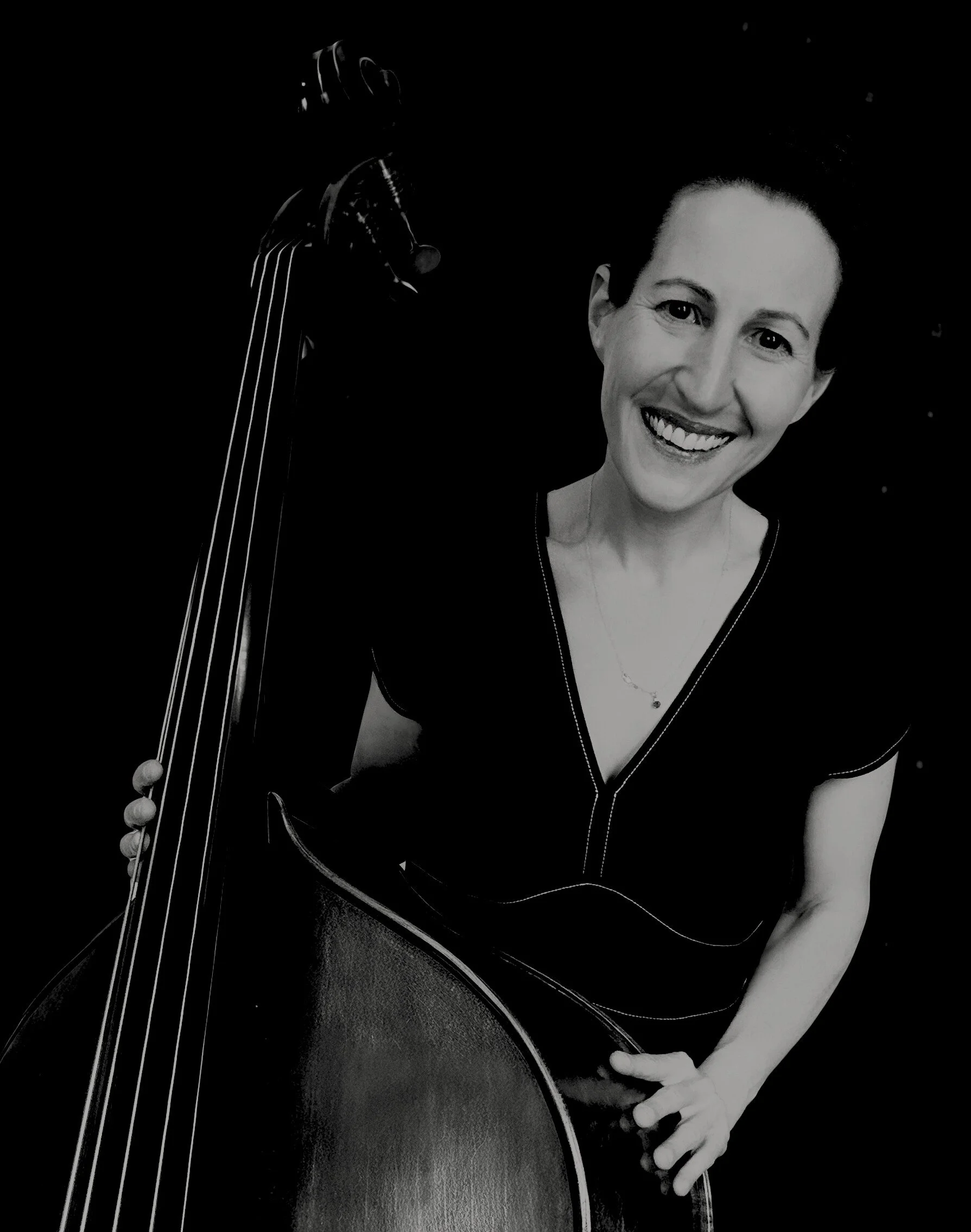 Black and white photo of a woman smiling, holding a bass guitar with her right hand, against a dark background.