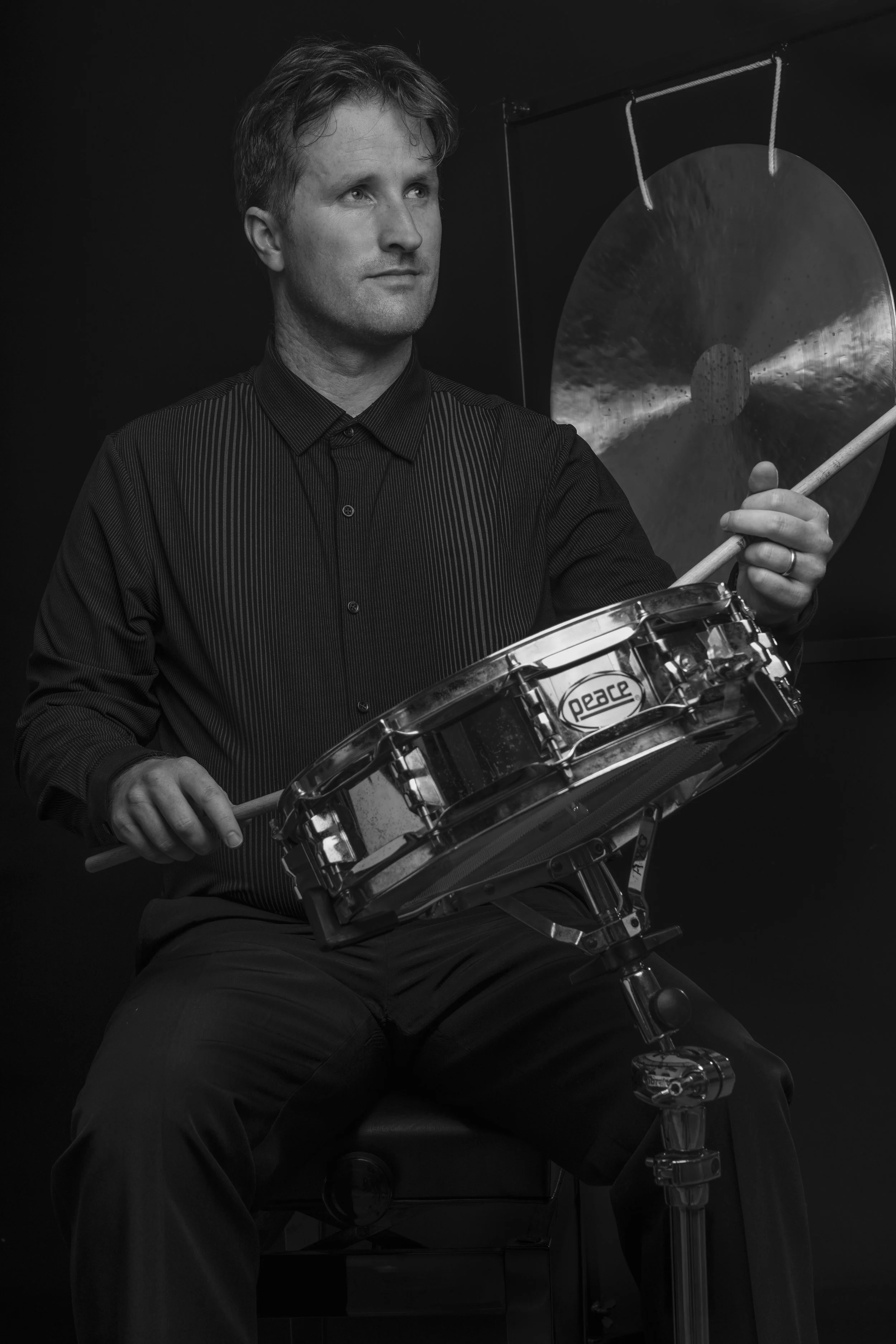 A man sitting while playing a snare drum, holding drumsticks, with a gong hanging in the background.