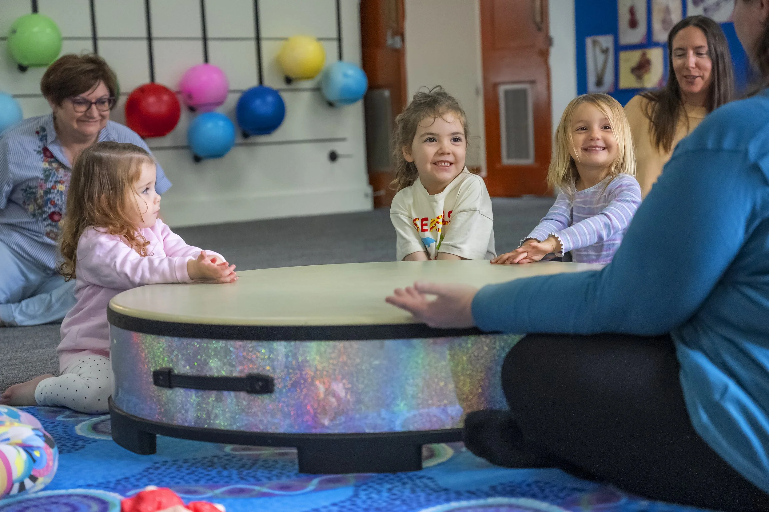 Group of young children with two adult women, sitting around a circular drum in a classroom with colorful balloons and educational posters on the walls.