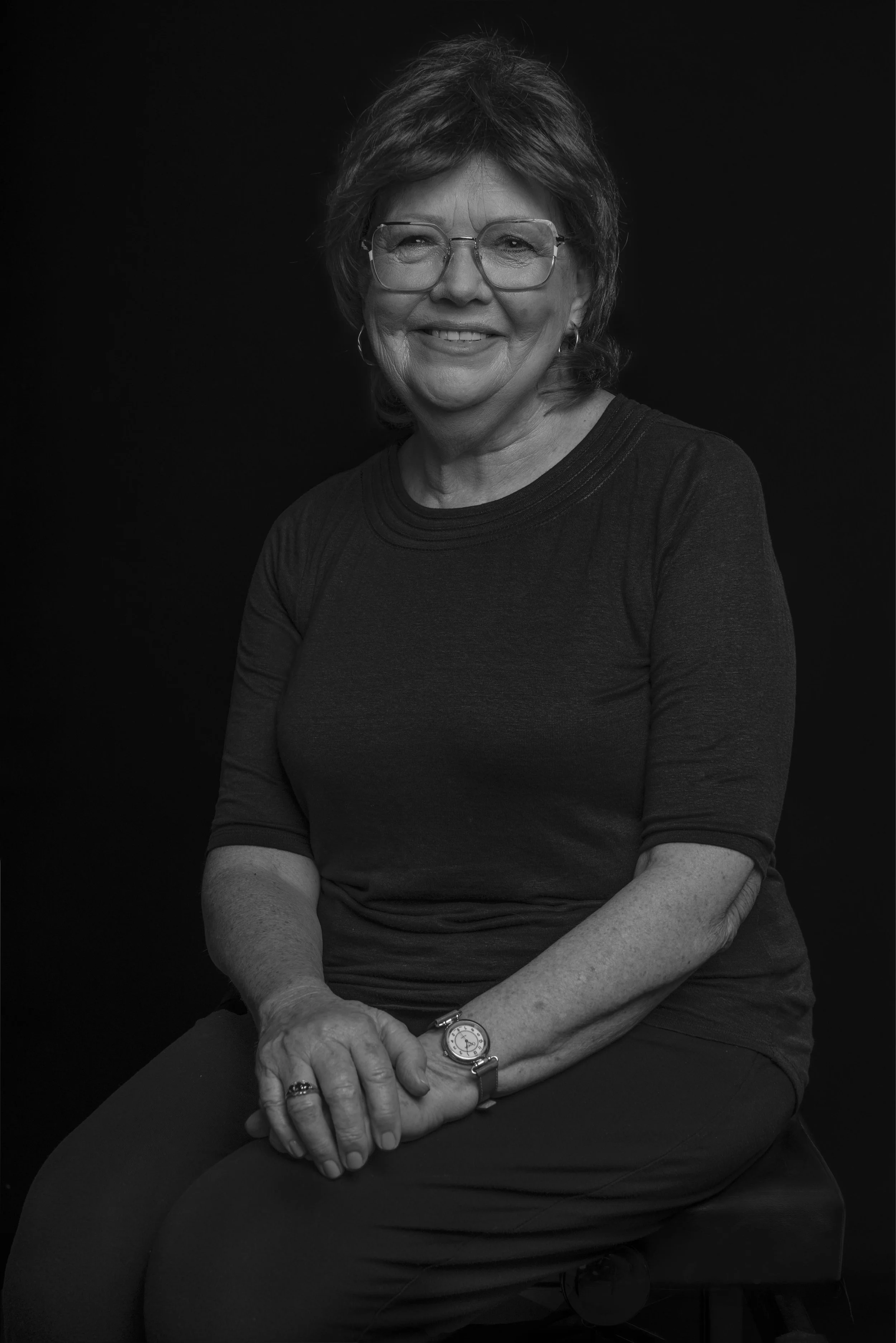 Black and white portrait of a woman with glasses and short hair, smiling and sitting on a stool against a dark background, wearing a dark shirt and pants.
