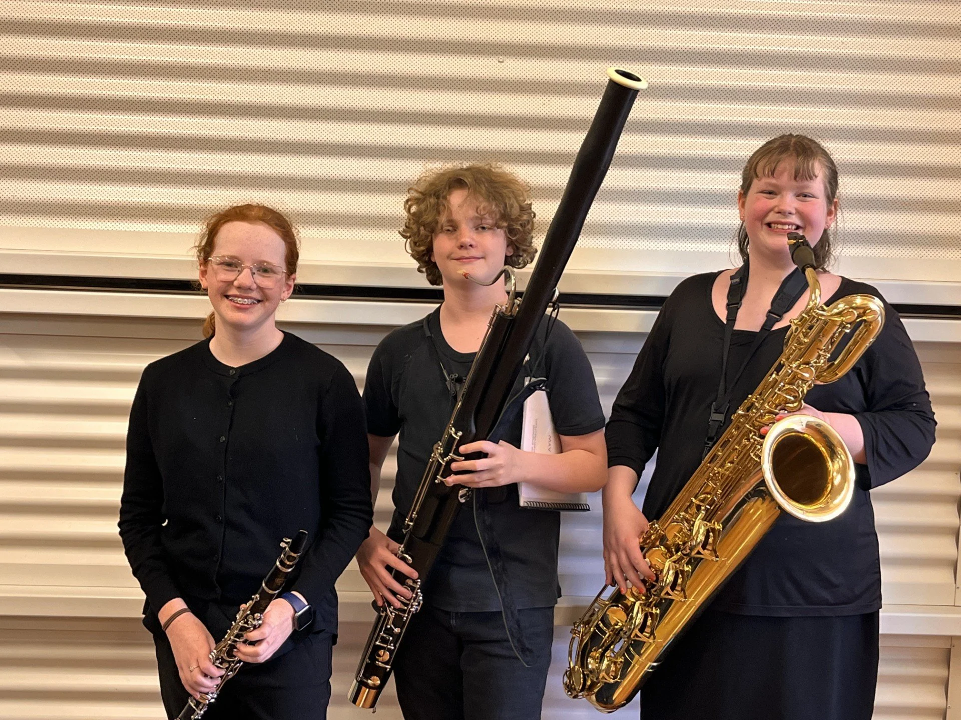 Three young girls standing side by side, holding musical instruments, and smiling. The girl on the left holds a clarinet, the girl in the middle holds a bassoon, and the girl on the right holds a saxophone.