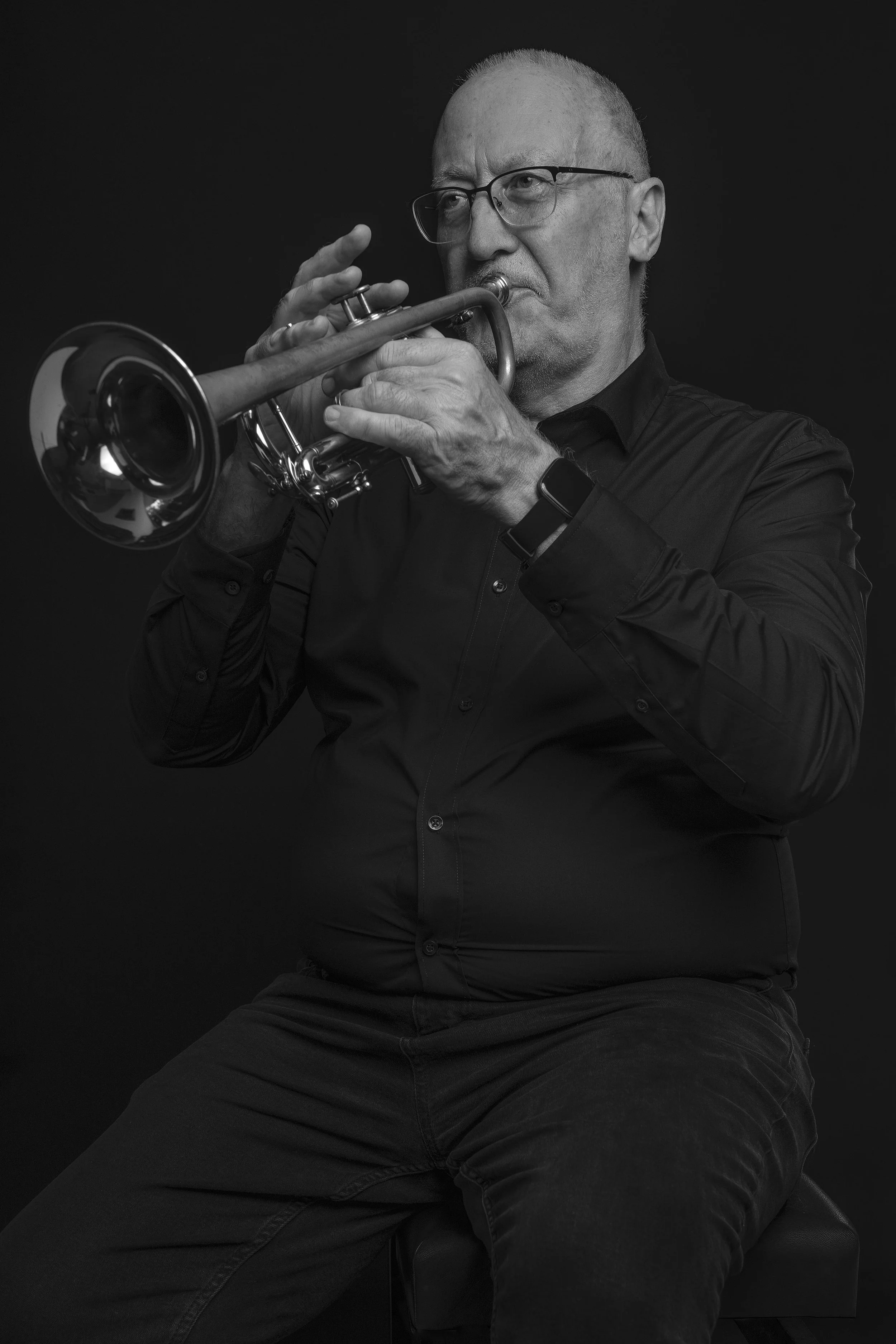 A black and white photo of a man with glasses, sitting on a stool playing a trumpet against a dark background.