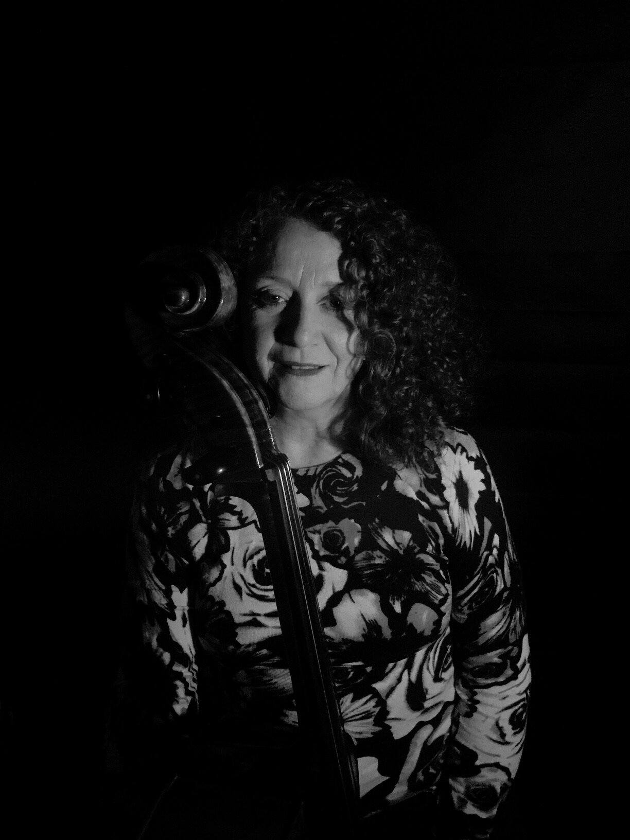 Black and white photo of a woman with curly hair, wearing a floral shirt, holding a cello, in low light.