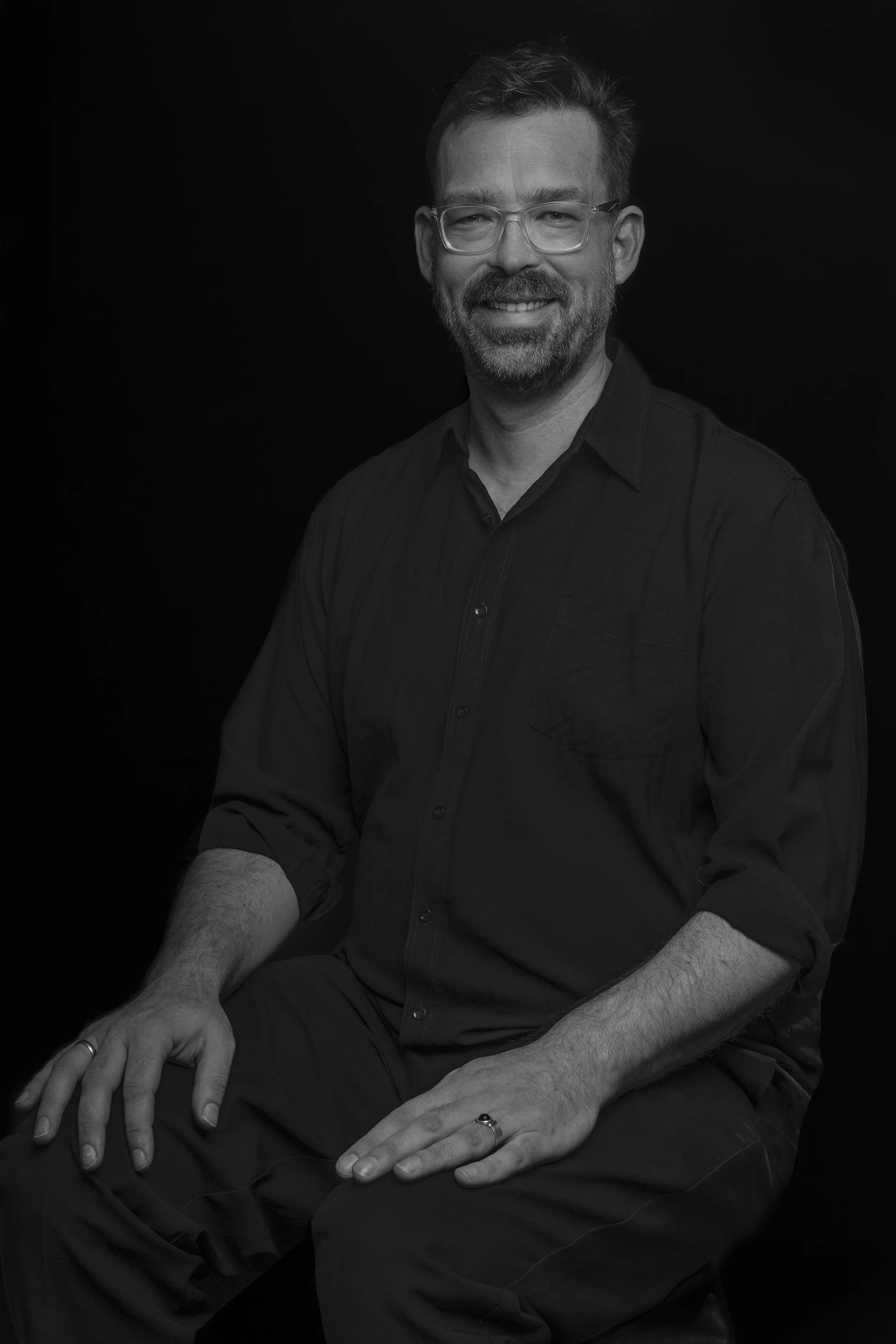 Black and white portrait of a smiling man with glasses, wearing a dark shirt, seated against a dark background.