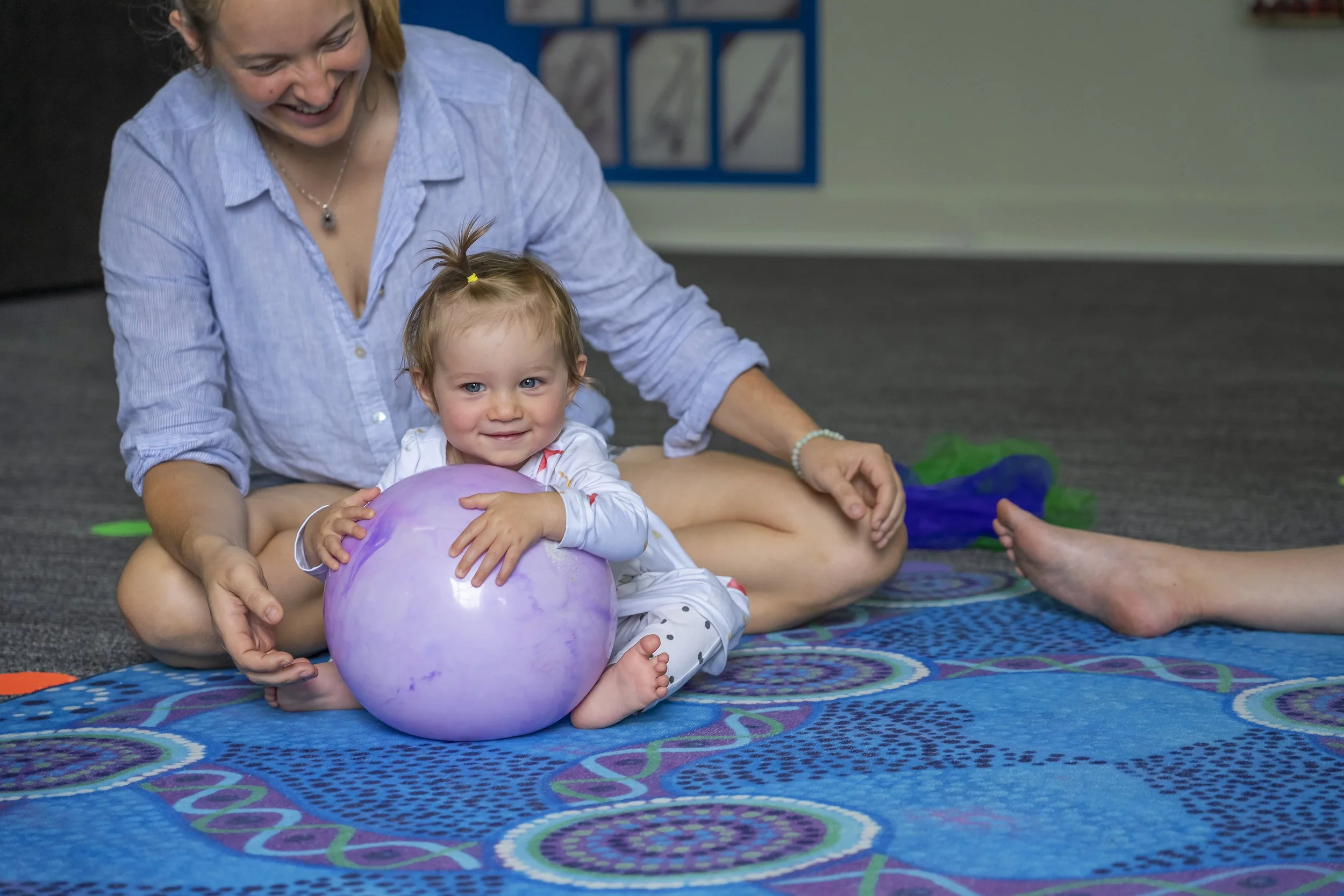 A woman and a young girl sitting on a colorful carpet. The girl is hugging a purple balloon and smiling, while the woman is smiling and sitting behind her. The girl has short, light brown hair with a small ponytail on top, and the woman has medium-length blonde hair.