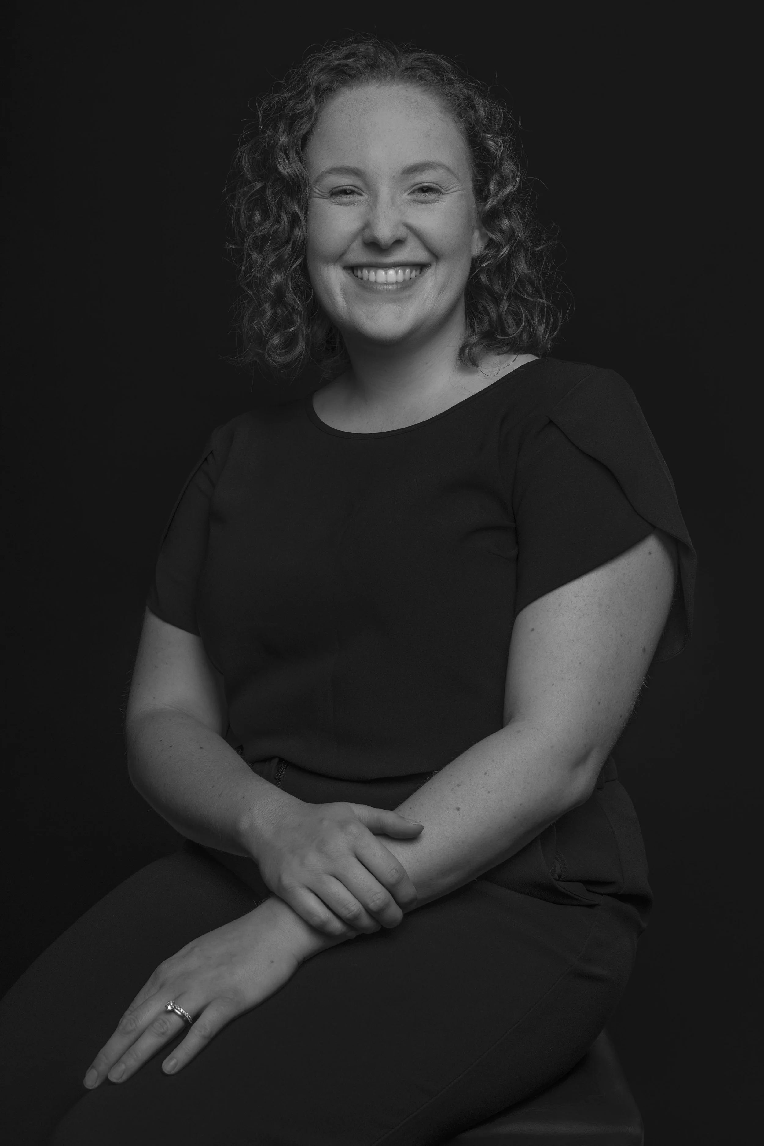 Black and white portrait of a woman smiling, with curly hair, wearing a black short-sleeved top, seated against a dark background.