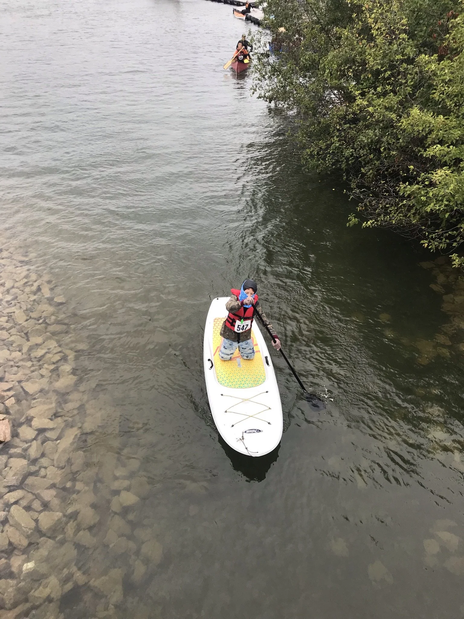 Child in a life jacket standing on a paddleboard, holding a paddle in the water. Other people in kayaks can be seen in the background.