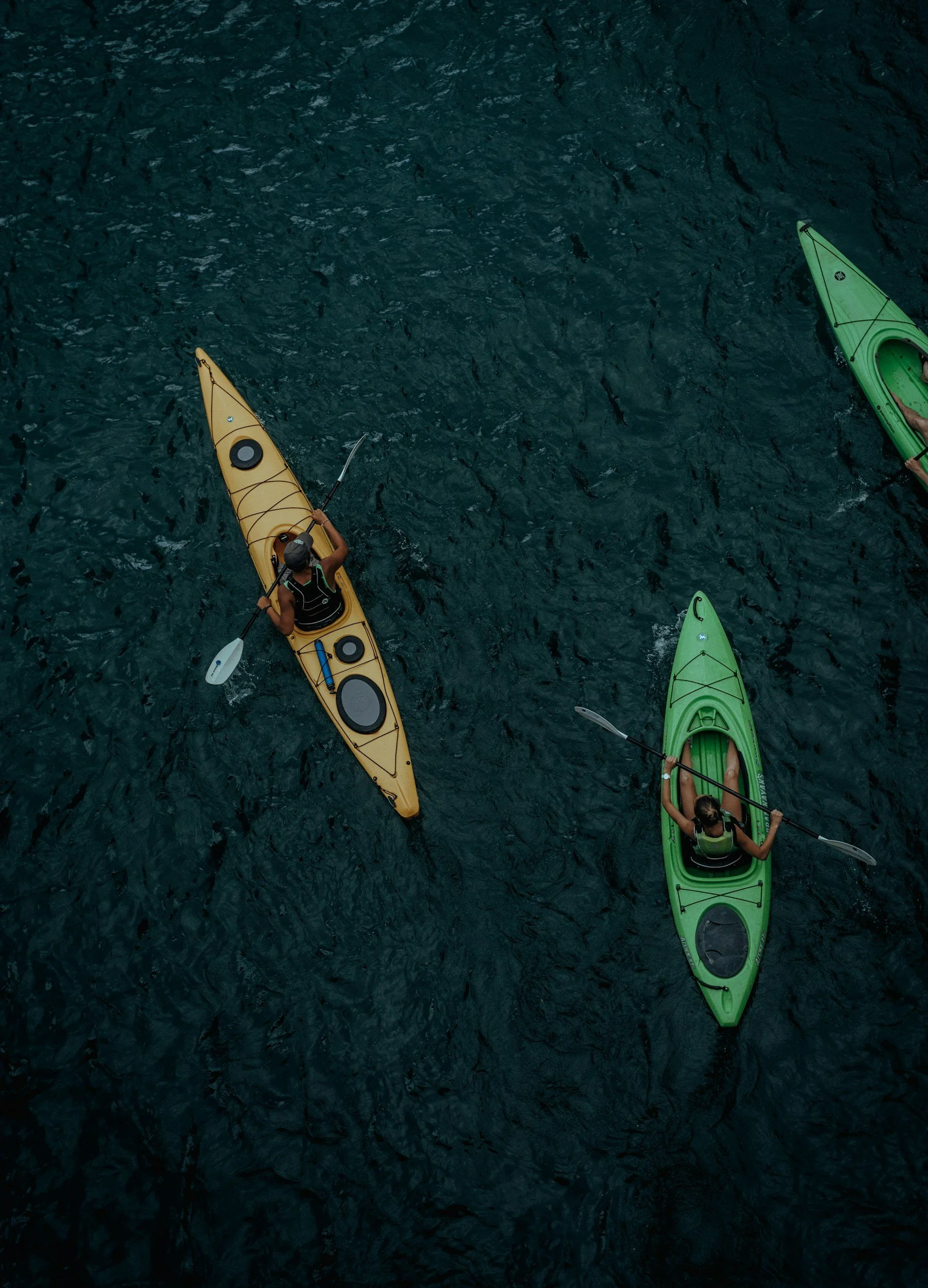 Two people kayaking on dark blue water, one in a yellow kayak and the other in a green kayak, viewed from above.