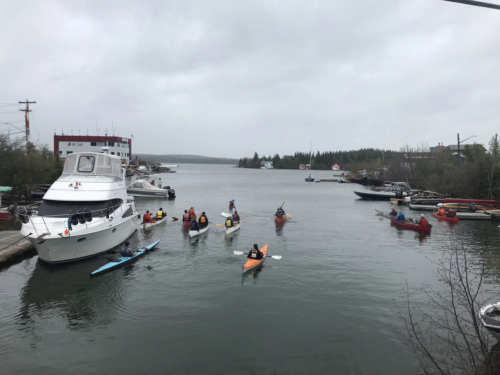People kayaking and paddleboarding in a bay with boats and a large building in the background on a cloudy day.