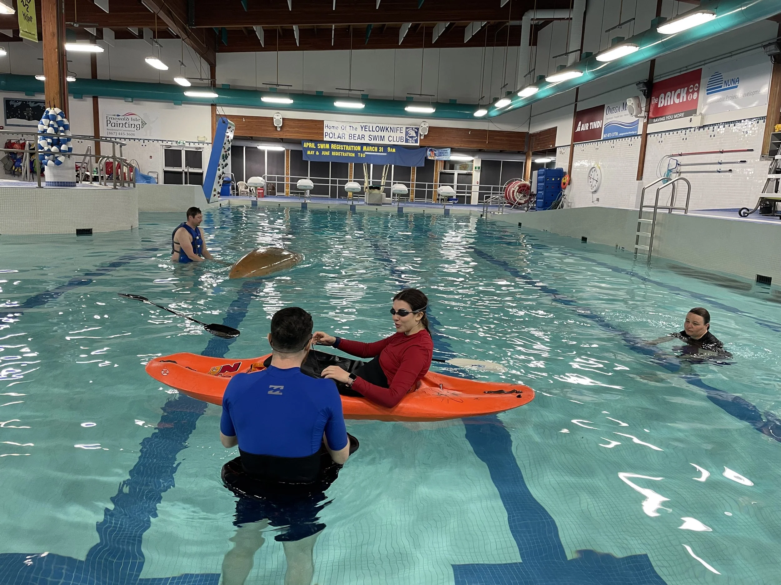 Indoor swimming pool with a person on a kayak being assisted by two instructors. 