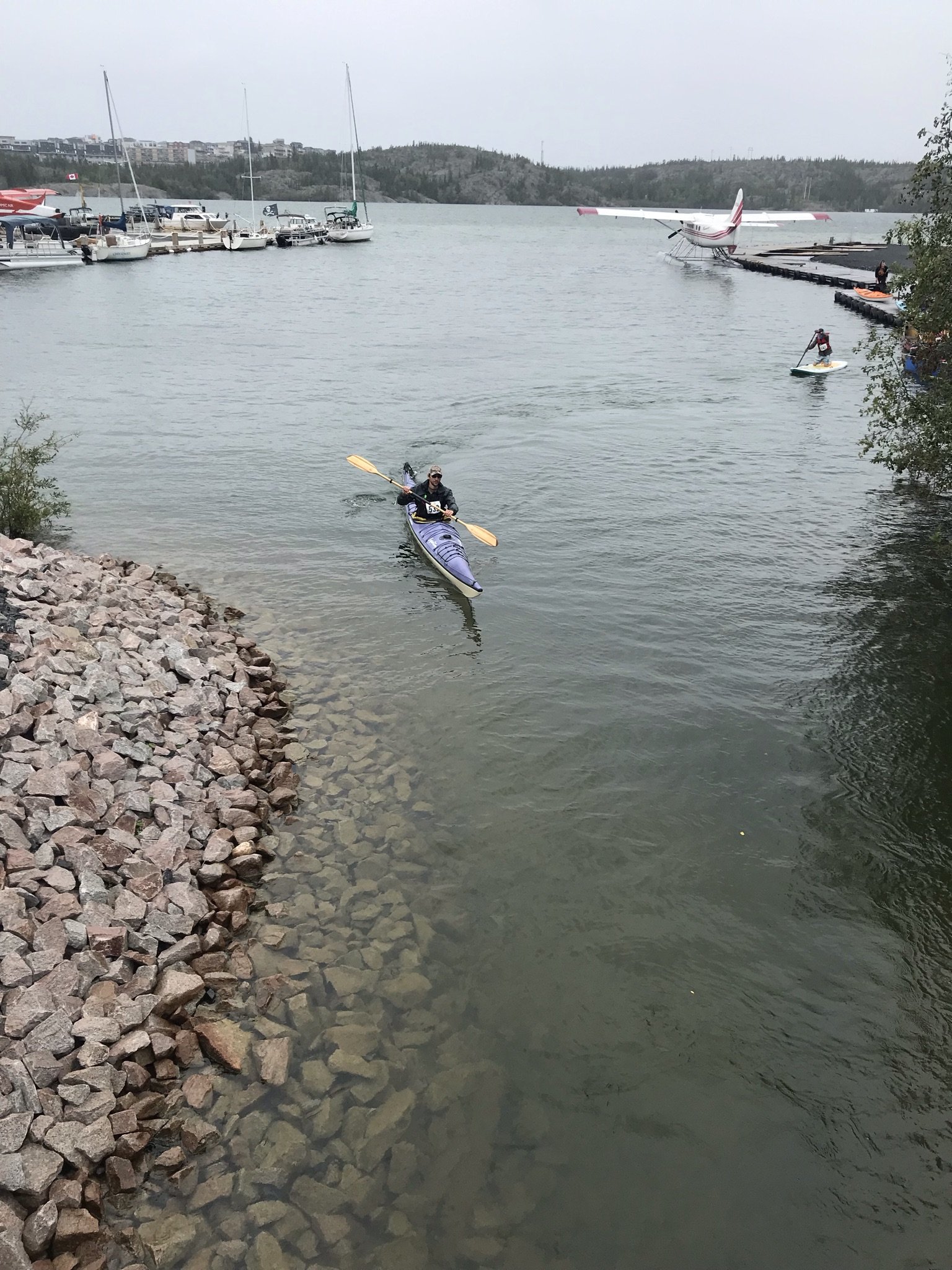A person kayaking near a rocky shoreline in a harbor with boats, sailboats, and a seaplane in the background.