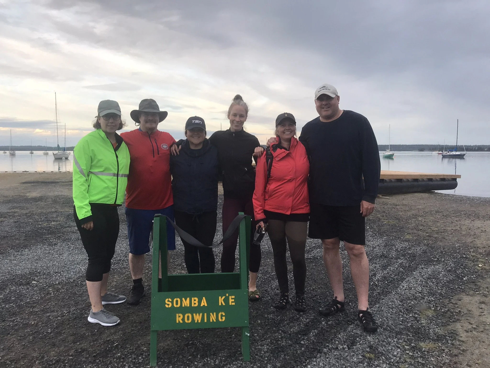 Group of six people standing outdoors near water with sailboats at rest in the background during sunset. They are dressed in athletic clothing, some with hats and sunglasses, and are smiling. A green sign with yellow text reads 'SOMBA KE ROWING' in f