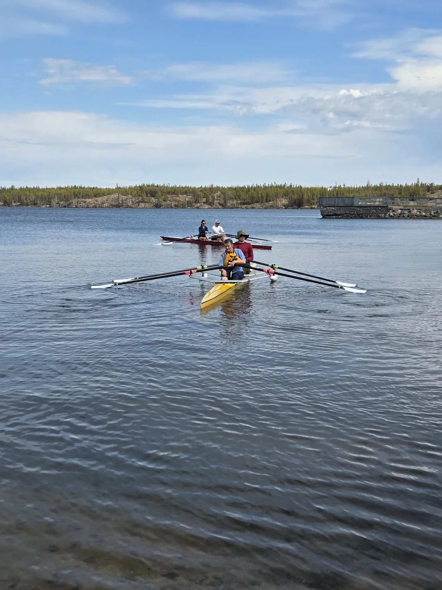 Three people are kayaking on a calm body of water under a partly cloudy sky, with a forested shoreline and a stone wall or dock in the background.