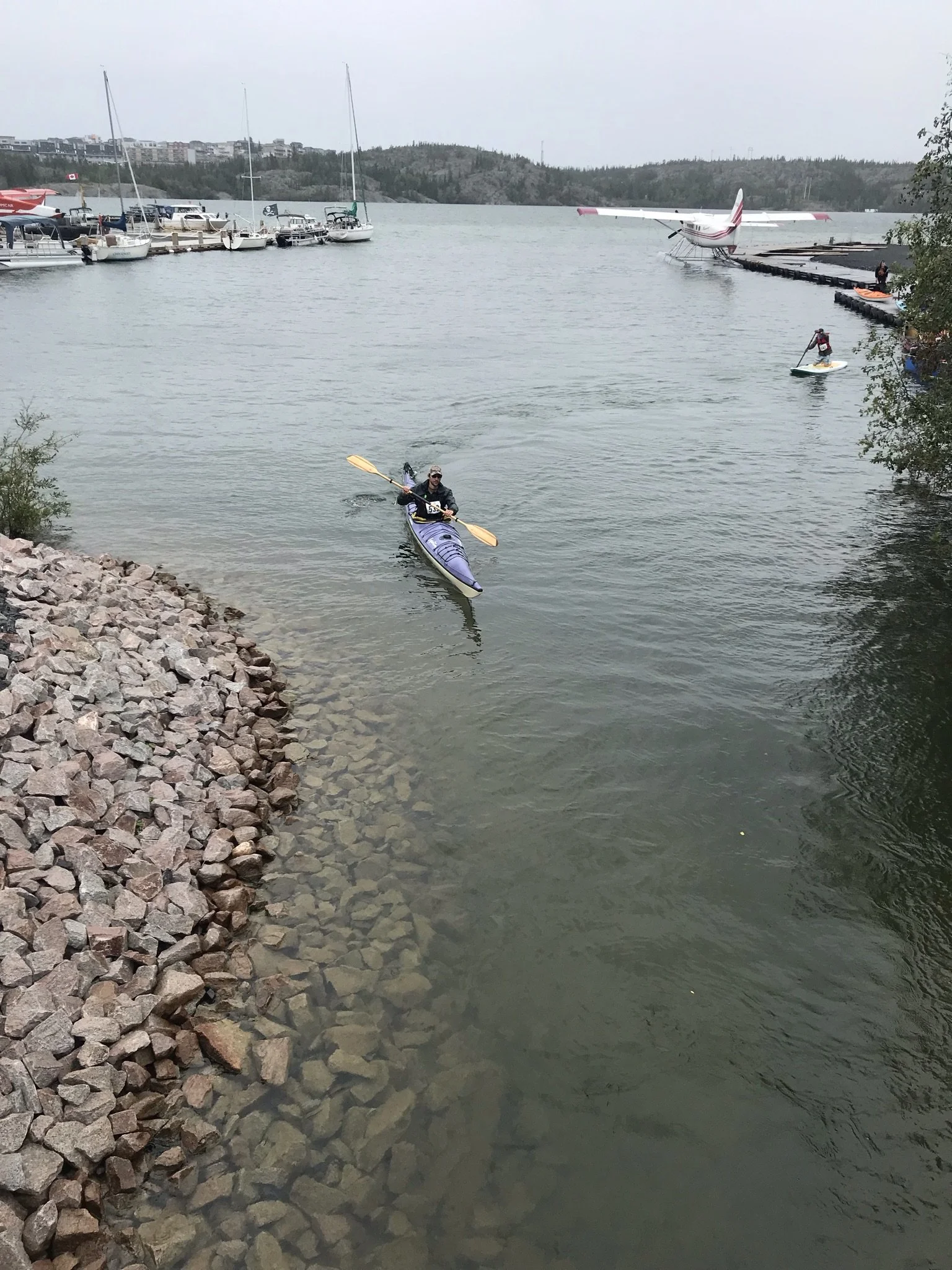 A person in a kayak paddling near the rocky shoreline of a marina, with boats docked and a small seaplane on the water in the background.