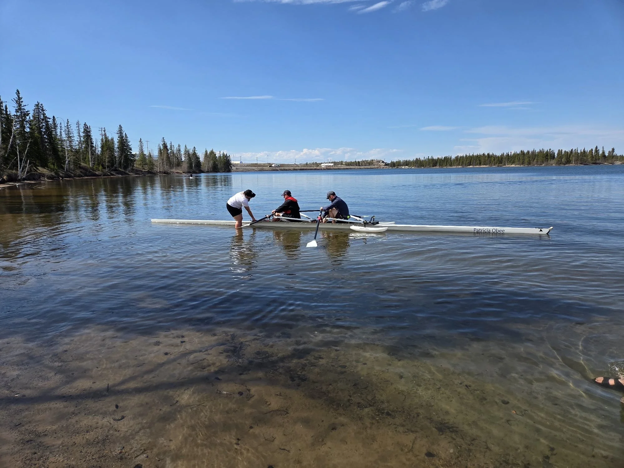 Three people preparing a kayak at the water's edge on a clear, sunny day, with trees on the shoreline and a blue sky.