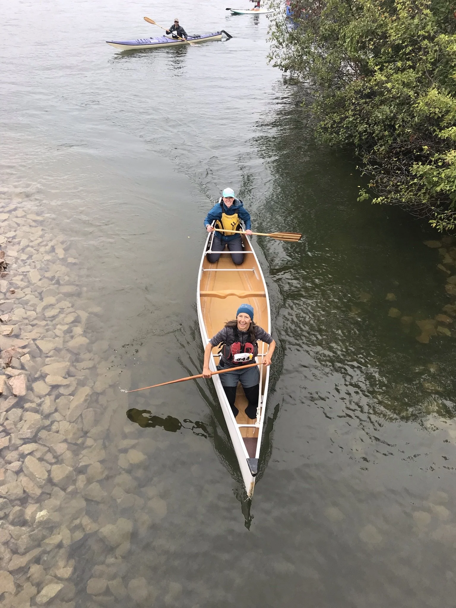 Two women smiling and paddling a kayak on a river, with a man in a kayak in the background near the riverbank covered with rocks and trees.