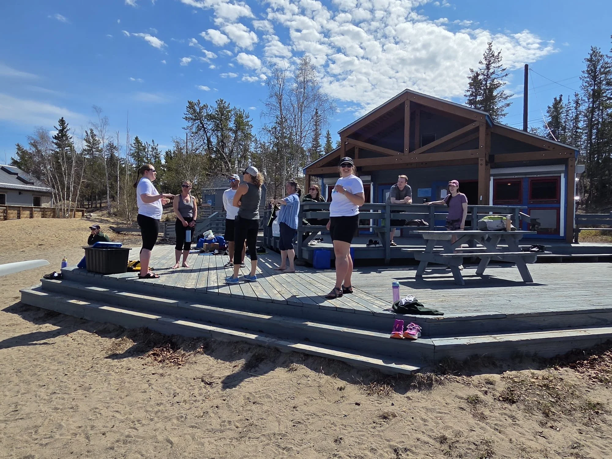 Group of people celebrating on a wooden deck outside a house, with some sitting on picnic tables, under a blue sky with scattered clouds, and trees in the background.