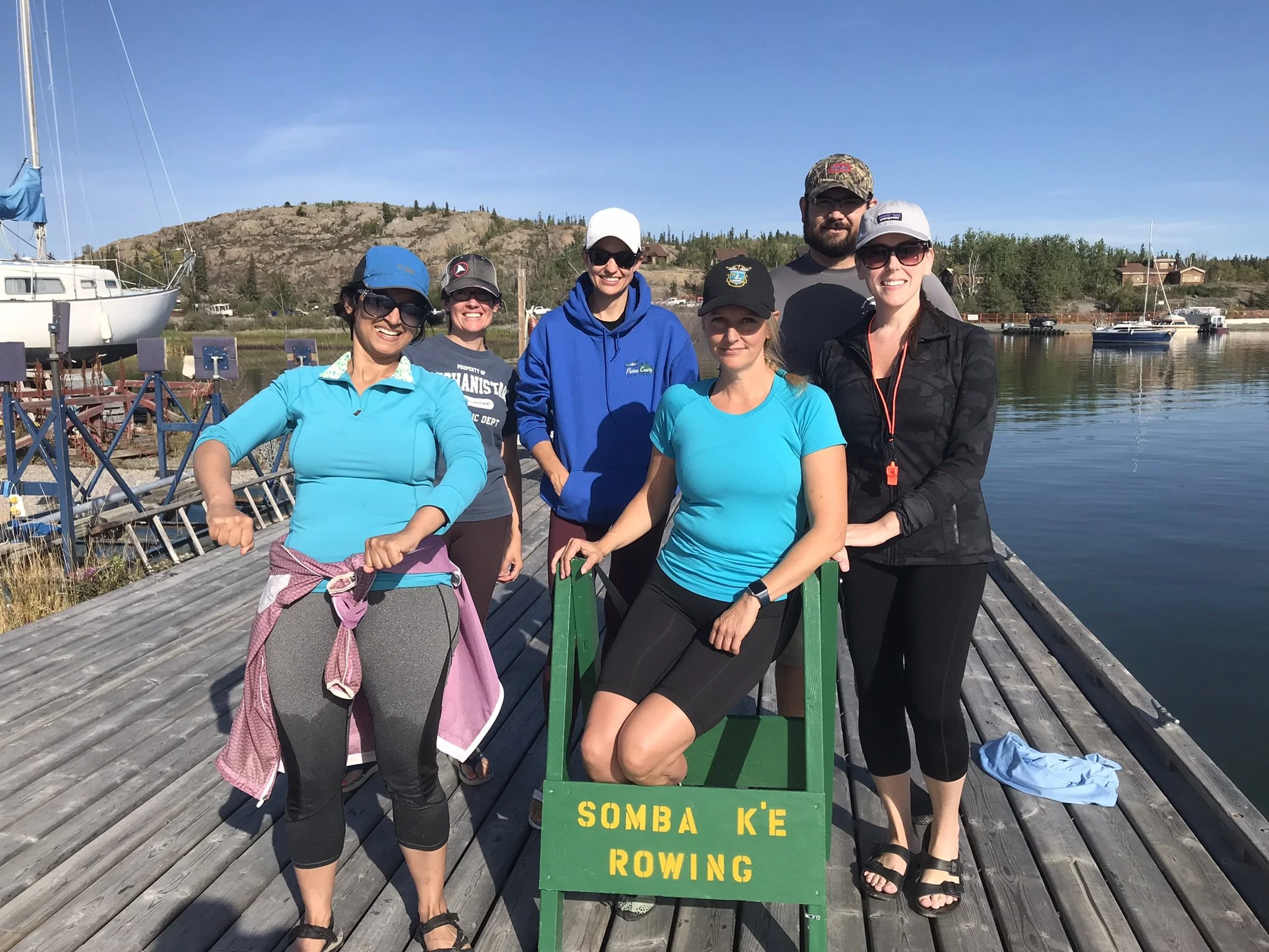 Group of six people standing on a wooden dock near boats and water, with a hillside and trees in the background, posing for a photo during daytime.
