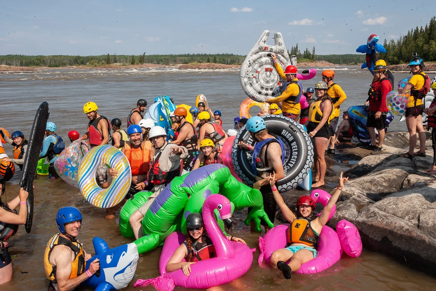 People wearing life jackets and helmets, holding and floating on inflatable pool toys like unicorns, flamingos, and donuts, gathered at the edge of a river on a sunny day.