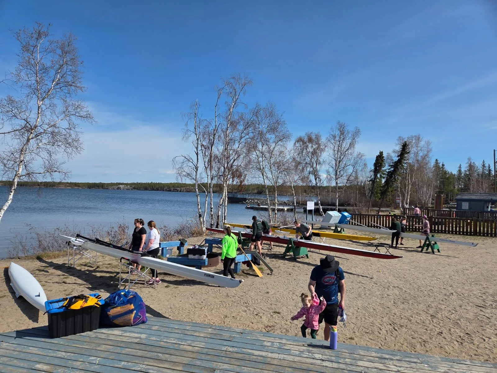 People preparing kayaks on the sandy shore of a calm lake with trees and a wooden fence in the background under a partly cloudy sky.