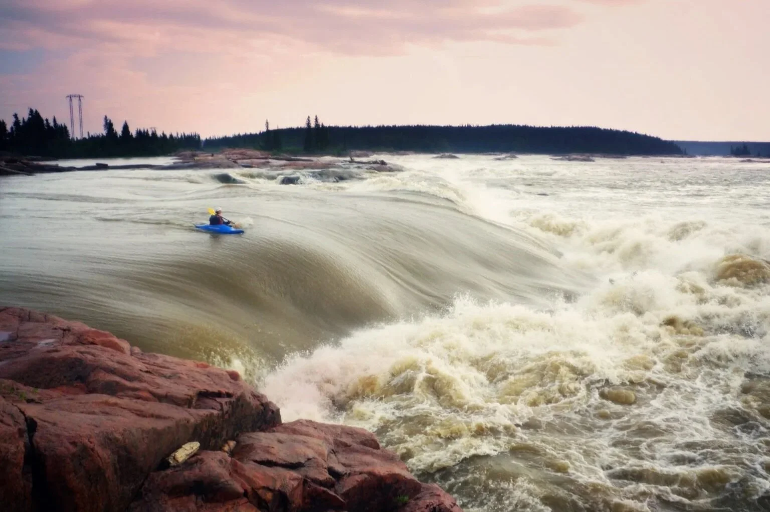 A kayaker paddling on a large, frothy river wave near rocky shoreline with trees and cloudy sky in background.