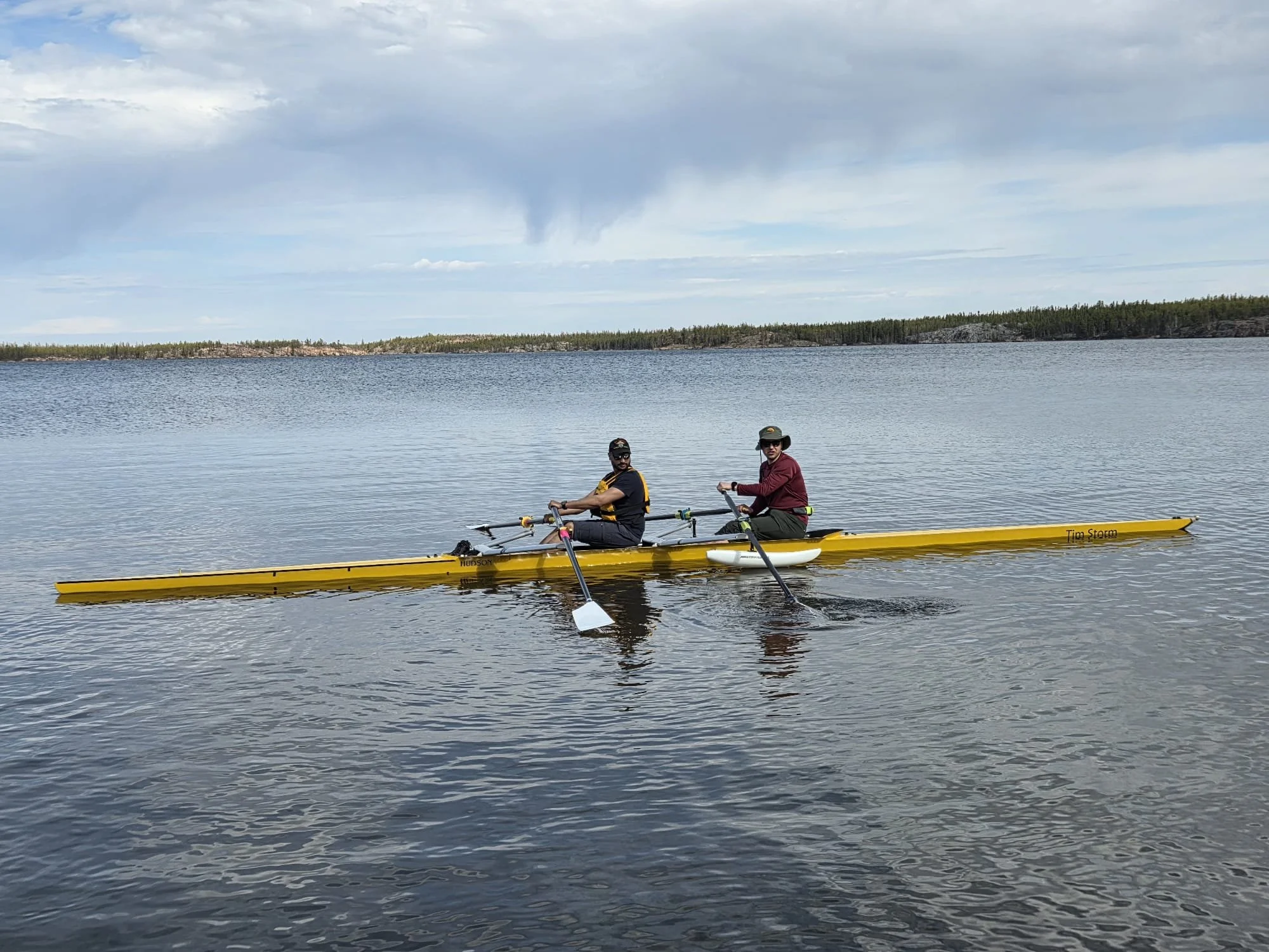 Two men kayaking on a calm lake with a distant tree-lined shoreline and cloudy sky.