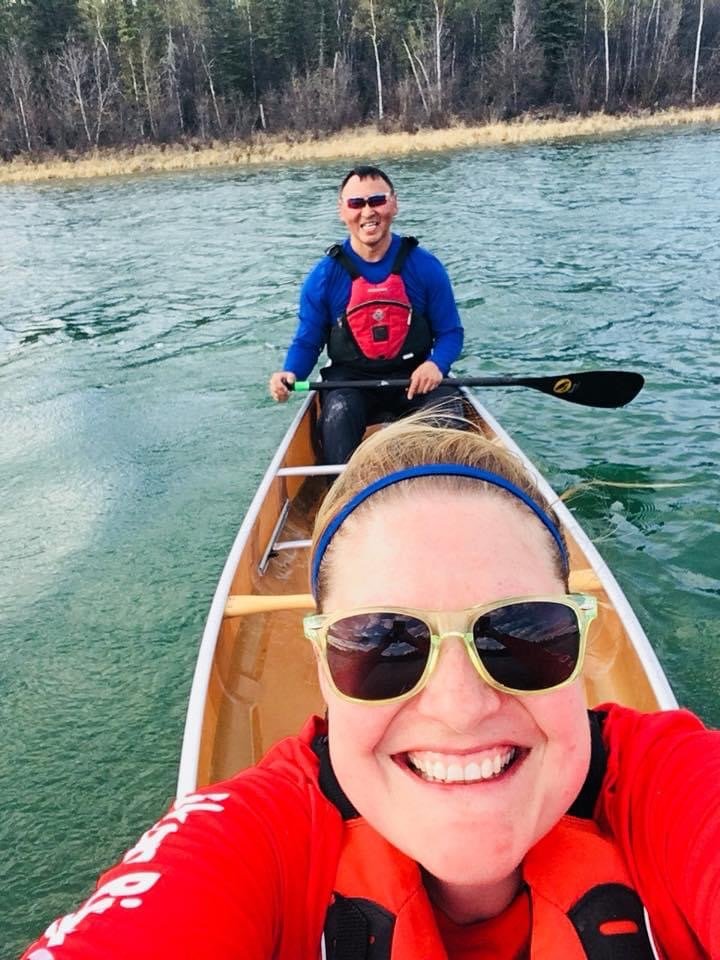Close-up of a smiling woman with curly afro hair, wearing glasses, hoop earrings, and a striped shirt, in an indoor setting.