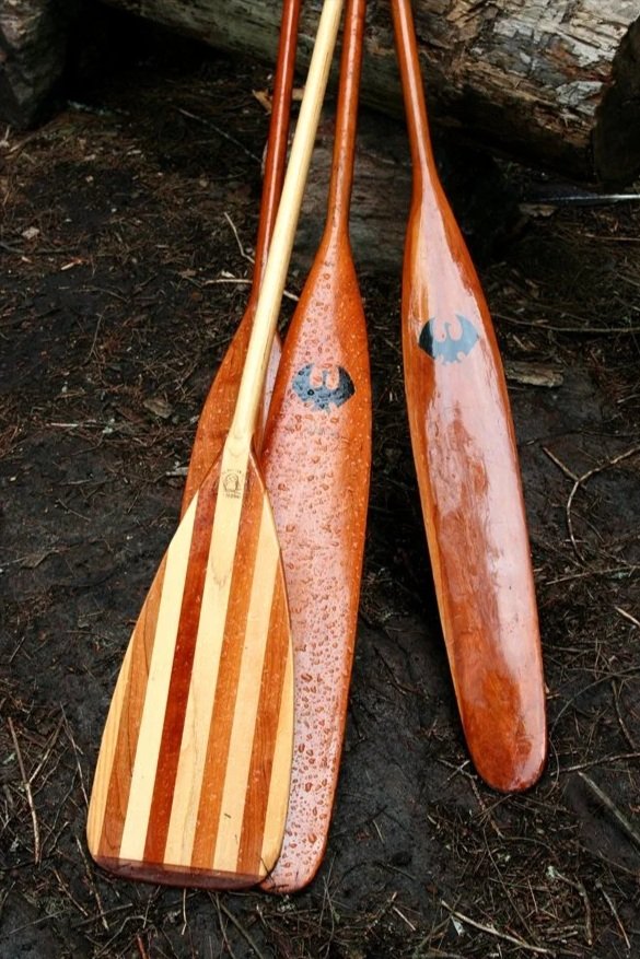 Three wooden paddles with one featuring a striped pattern and two with a solid finish, resting on dark dirt ground outdoors.