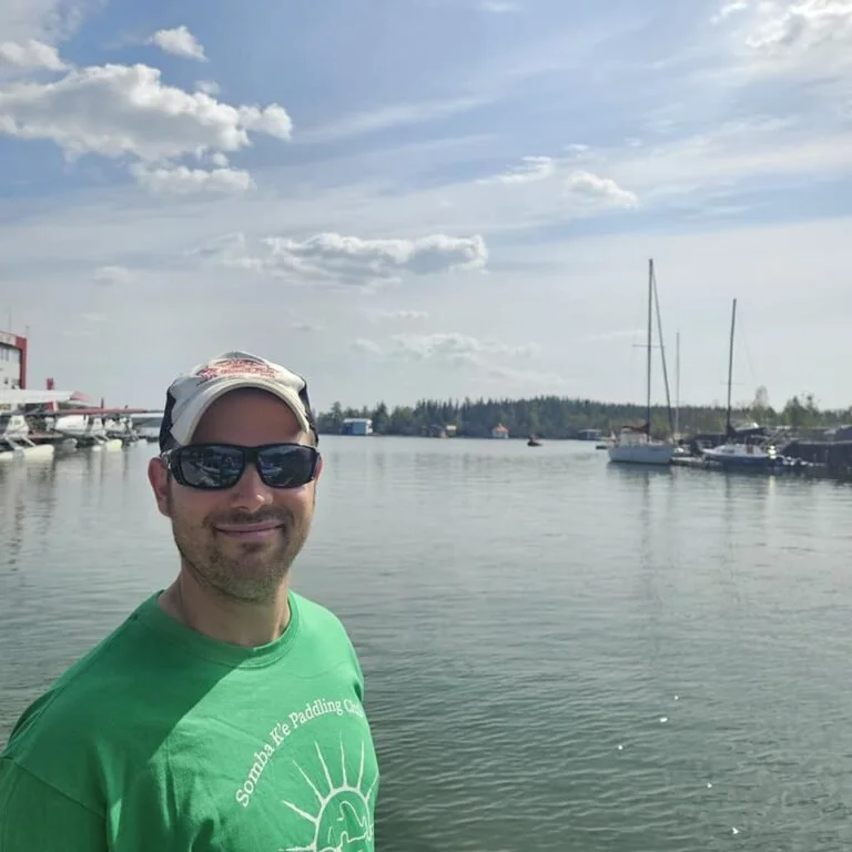 Smiling young woman with shoulder-length black hair, wearing a blue top, standing among green plants in a bright indoor setting.
