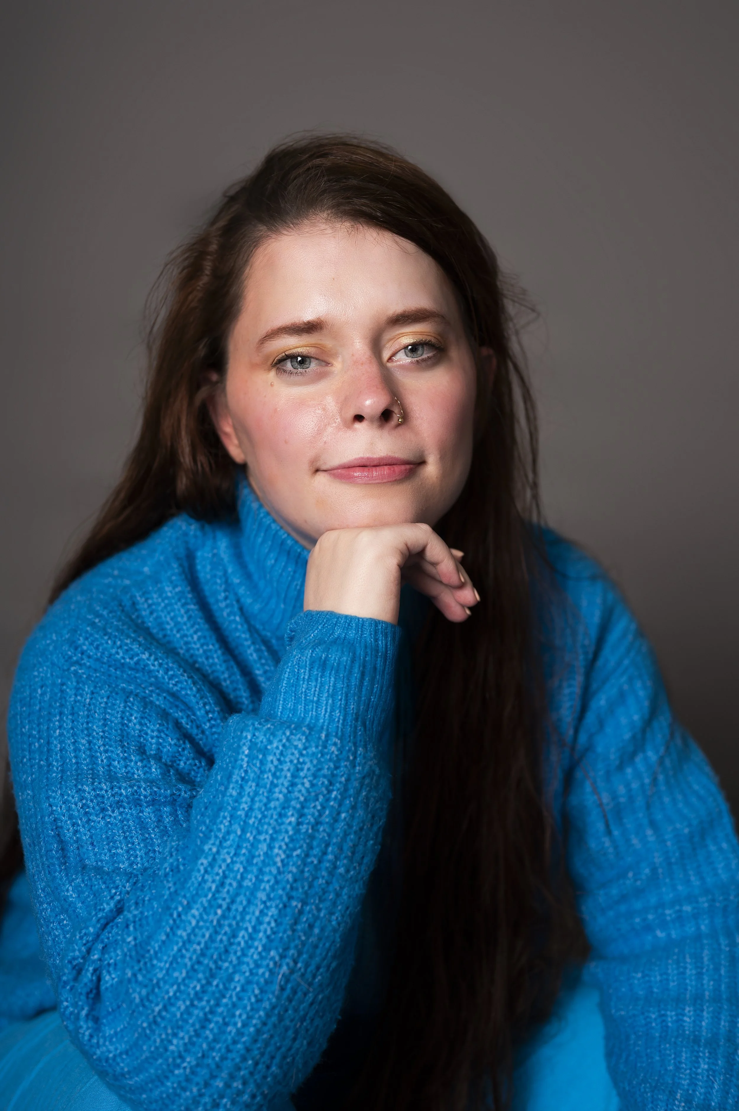 Elsa avec un pull bleu, du maquillage doré, les cheveux libres et un anneau de nez. Main positionnée sous le menton