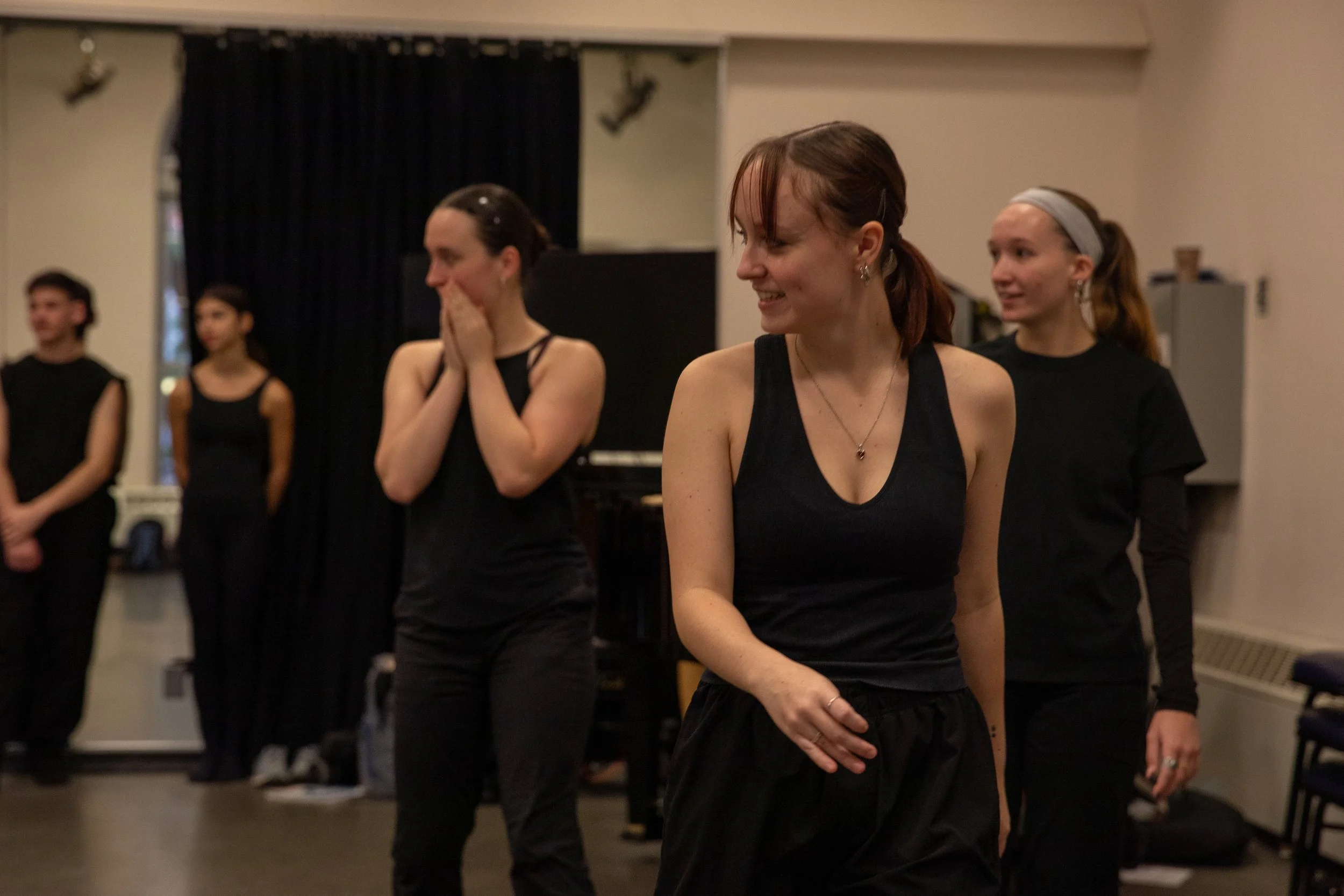 Group of five women practicing dance in a studio, dressed in black workout clothes, some smiling and others focused.