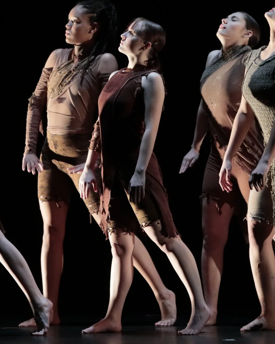 Group of dancers performing on stage, wearing earthy-toned costumes, with some dancers having their eyes closed and bodies positioned expressively.