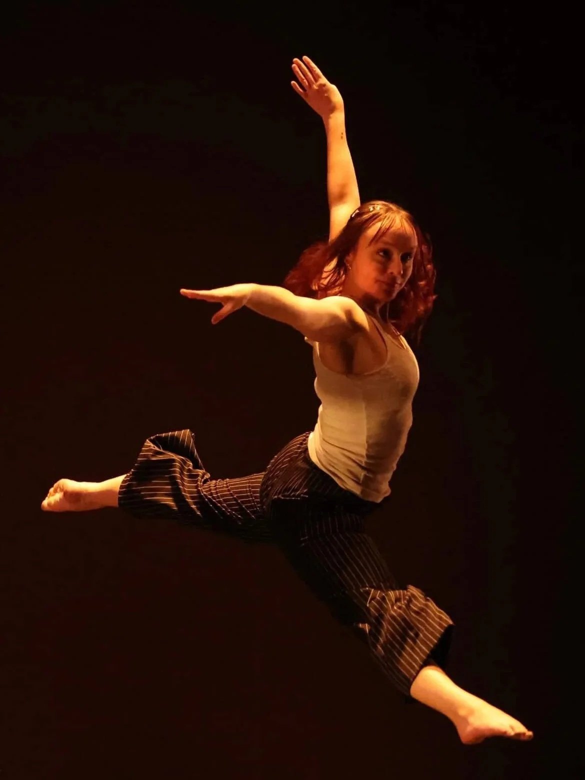 A woman with red hair performing a leap in a dance pose against a dark background, wearing a beige top and black pants with a striped pattern.
