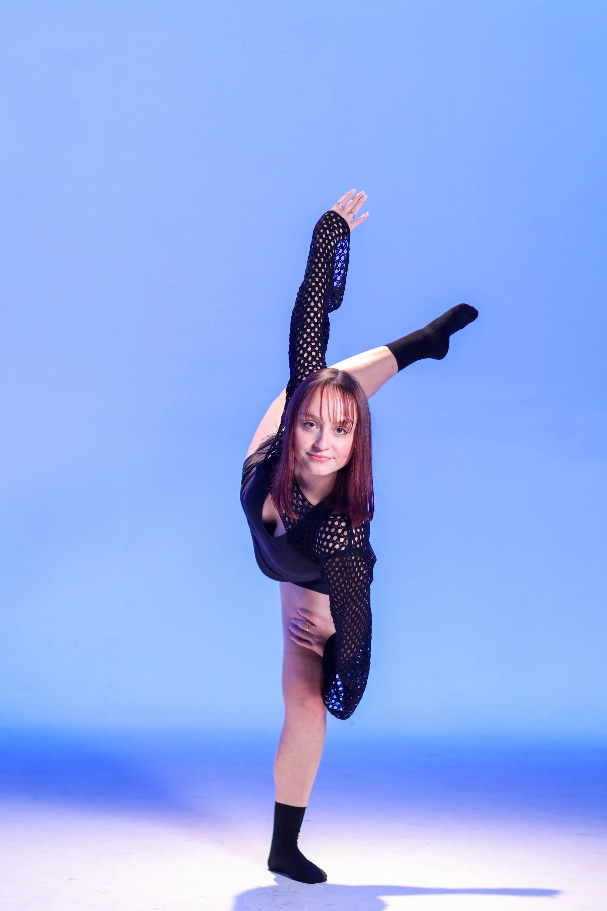 Woman doing a standing split dance pose against a blue background, wearing a black outfit with mesh sleeves and socks.