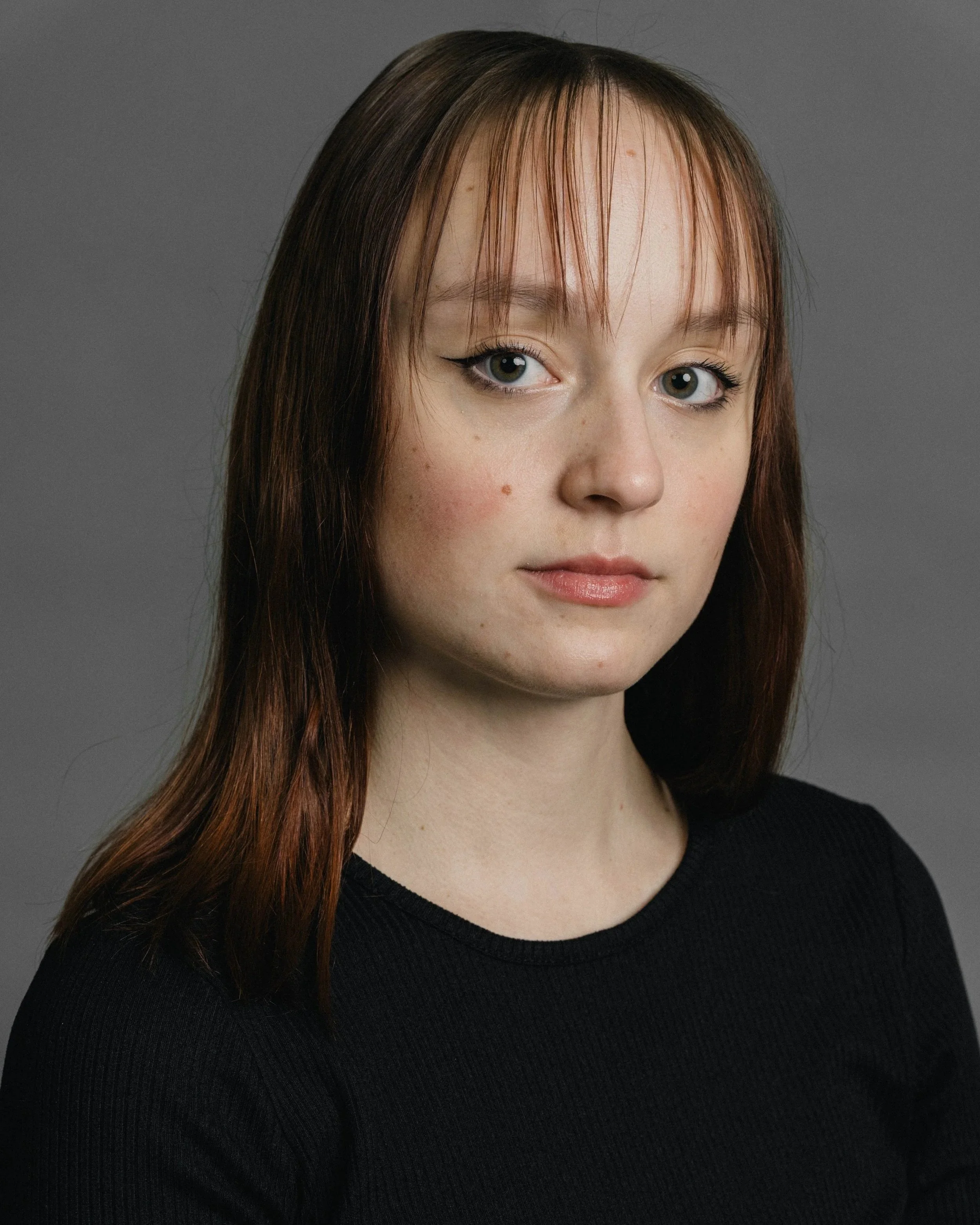 Portrait of a young woman with straight, shoulder-length brown hair, freckles, and light makeup, wearing a black top, against a gray background.