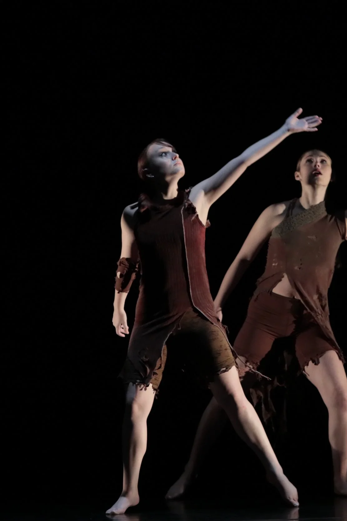 Two dancers on stage, one reaching upward with her arm, wearing torn brown costumes, against a dark background.