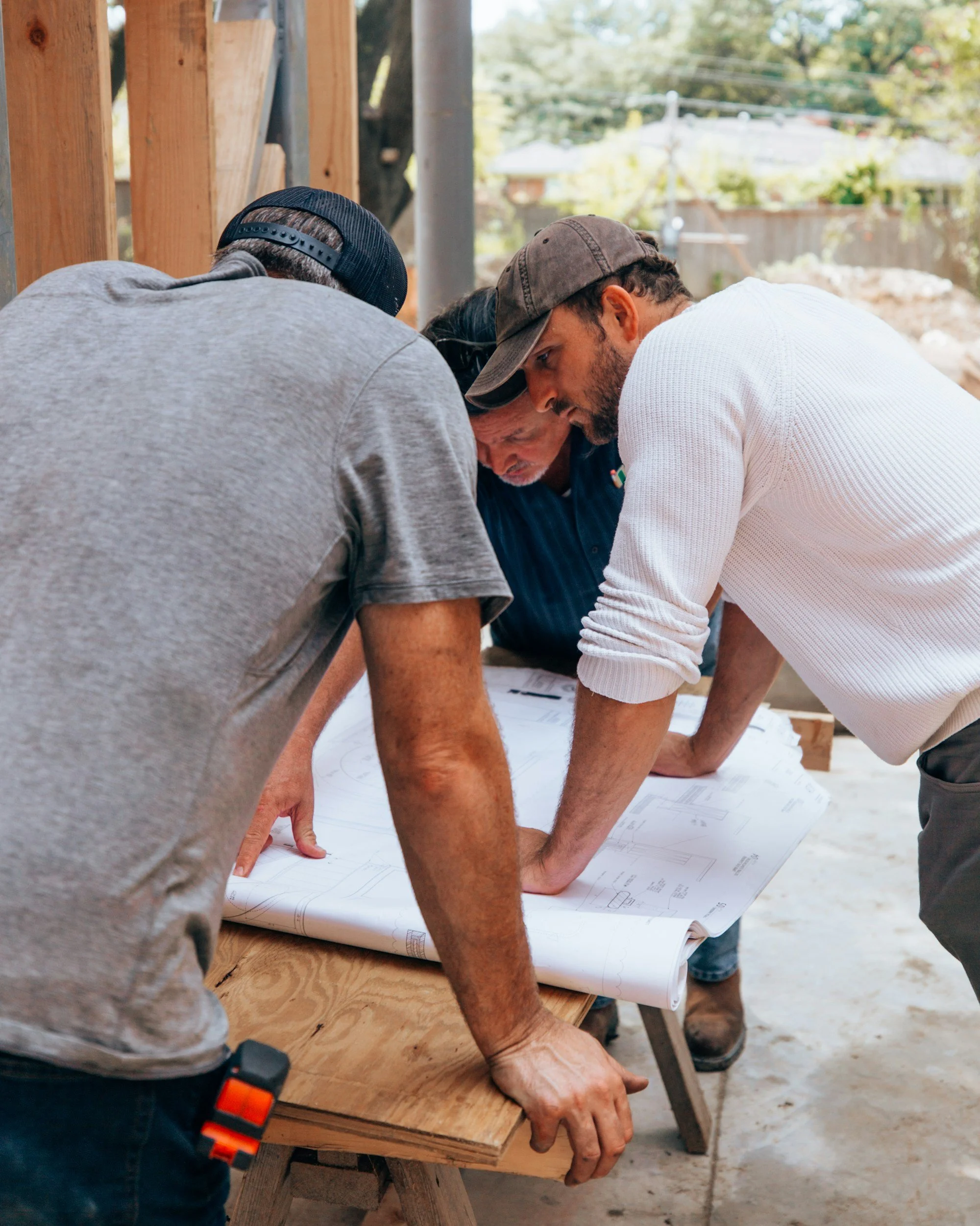 Three men at a construction site reviewing blueprints on a wooden table.