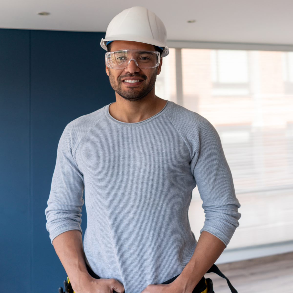 Construction worker smiling indoors, wearing a white hard hat and safety glasses, in a modern building with large windows.
