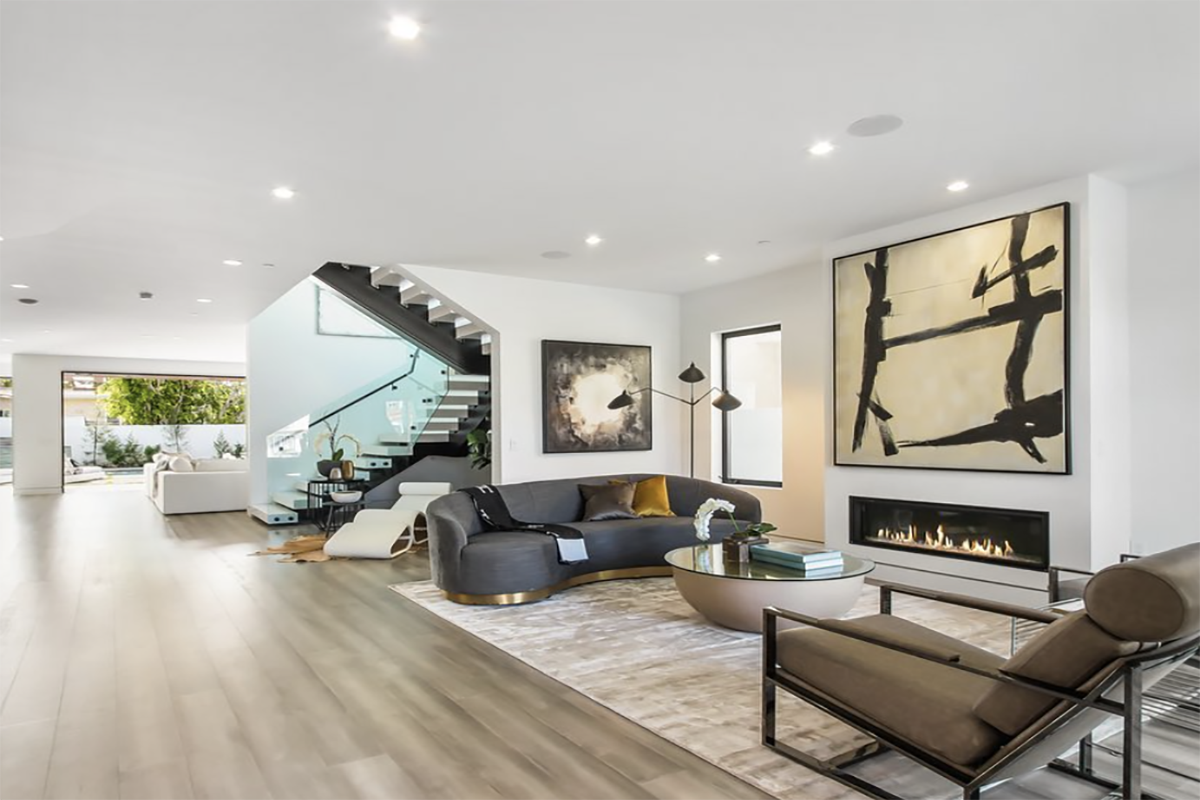Modern living room with a dark gray curved sofa, abstract art on white walls, a gas fireplace, and a glass staircase with a black handrail.