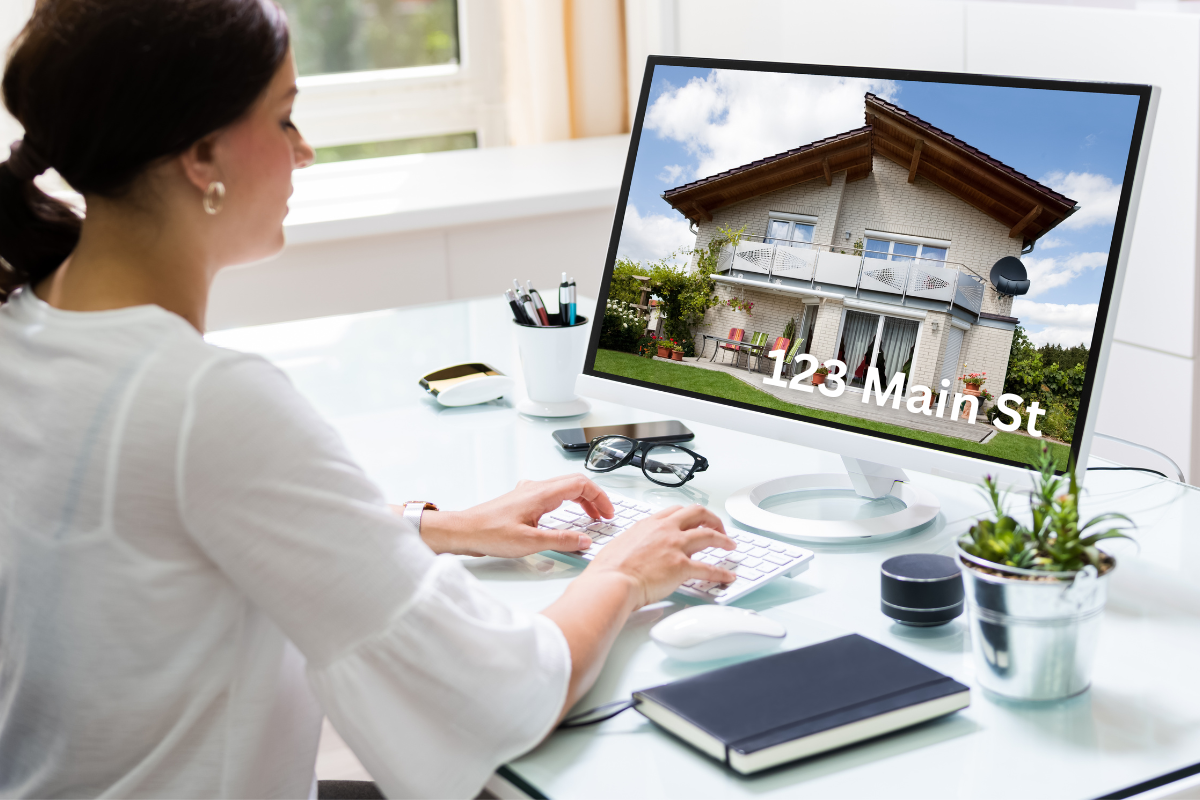 Woman using a computer at a desk, with a house and address on the monitor screen.