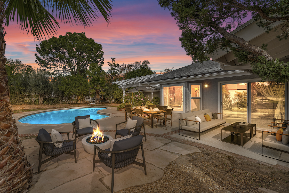 A backyard at sunset with a swimming pool, outdoor fire pit with chairs, dining table, and outdoor seating area with sofas and a coffee table.