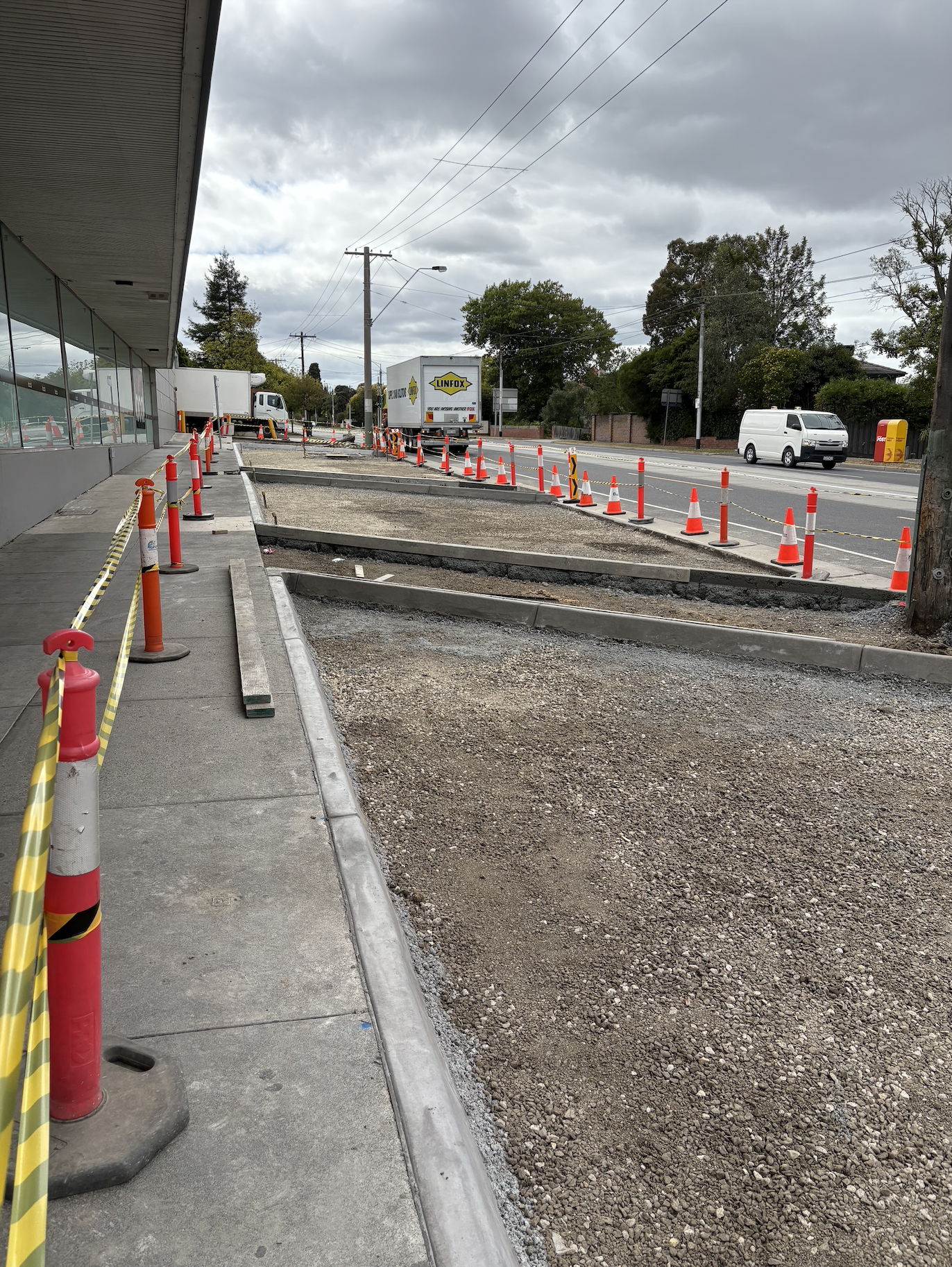 Construction site on a sidewalk with traffic cones, barriers, and construction equipment, with a road and moving vehicles in the background.