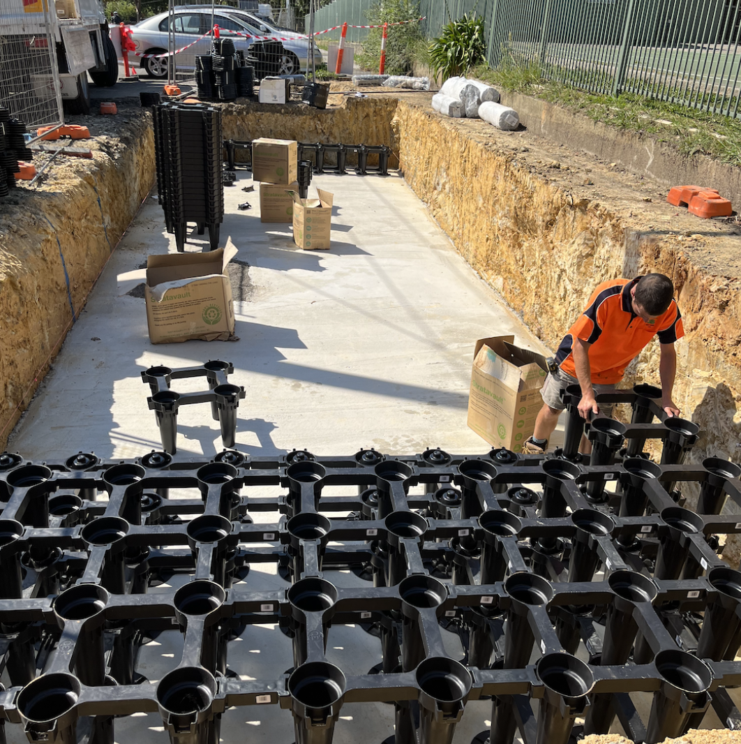 Construction worker installing a prefabricated water barrier system in a trench. Several black modular units are stacked nearby on a concrete surface, with other construction materials and equipment around.