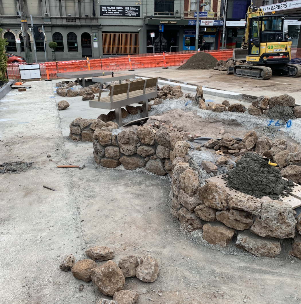 Construction site with rocks and dirt, partially built stone wall, machinery, and orange safety barriers in an urban area.