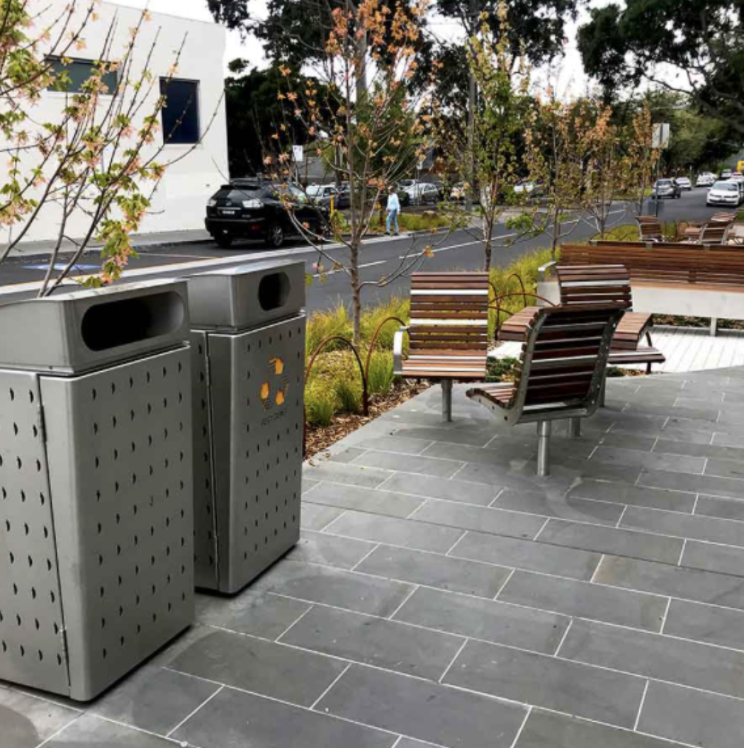 Empty outdoor seating area with wooden benches, trees with sparse leaves, and two metal trash bins on a tiled sidewalk, with a street and cars in the background.