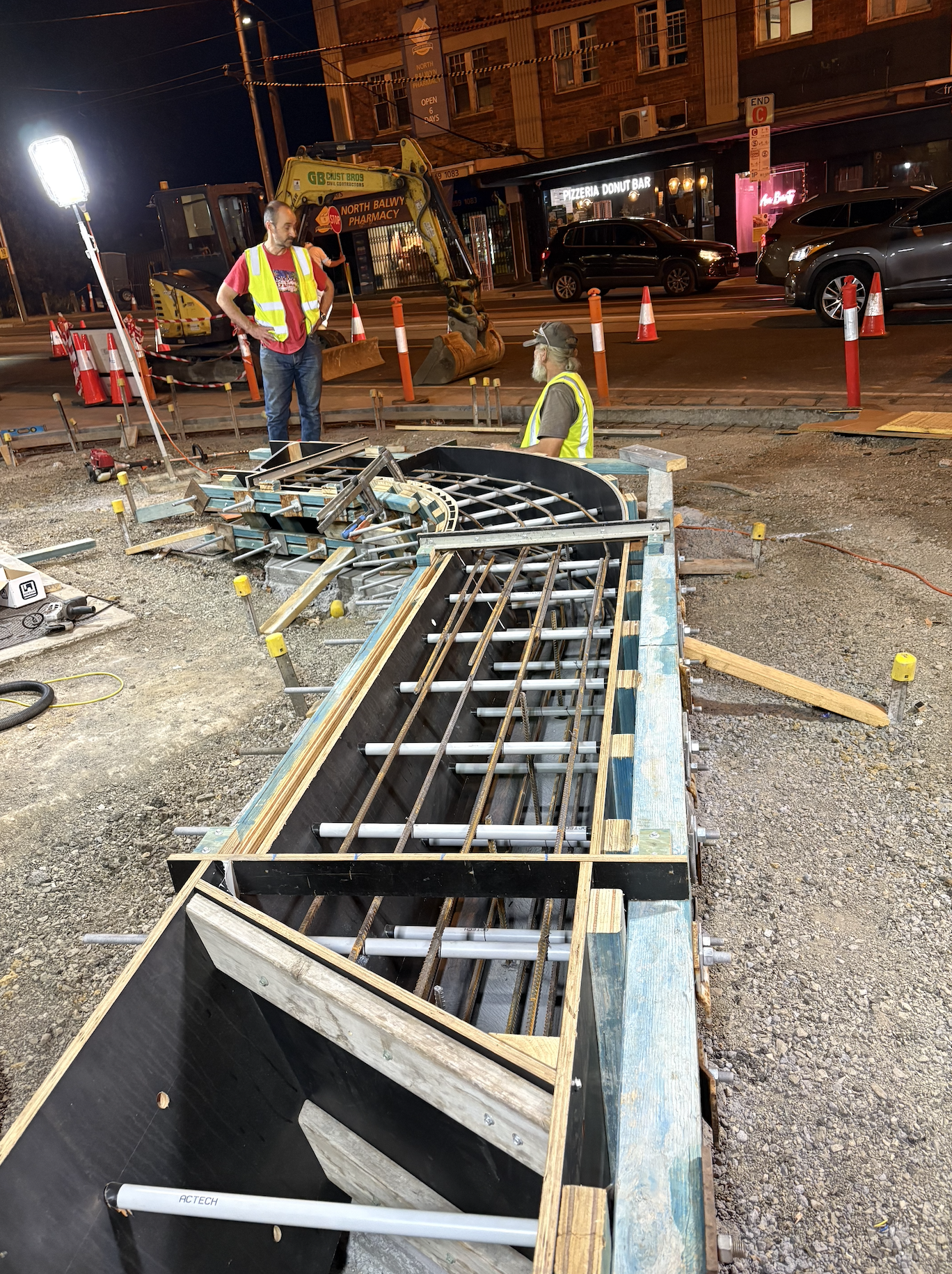 Construction workers at night working on a road with construction equipment and barriers, talking near a large trench with rebar and pipe framework, with cars and a storefront in the background.