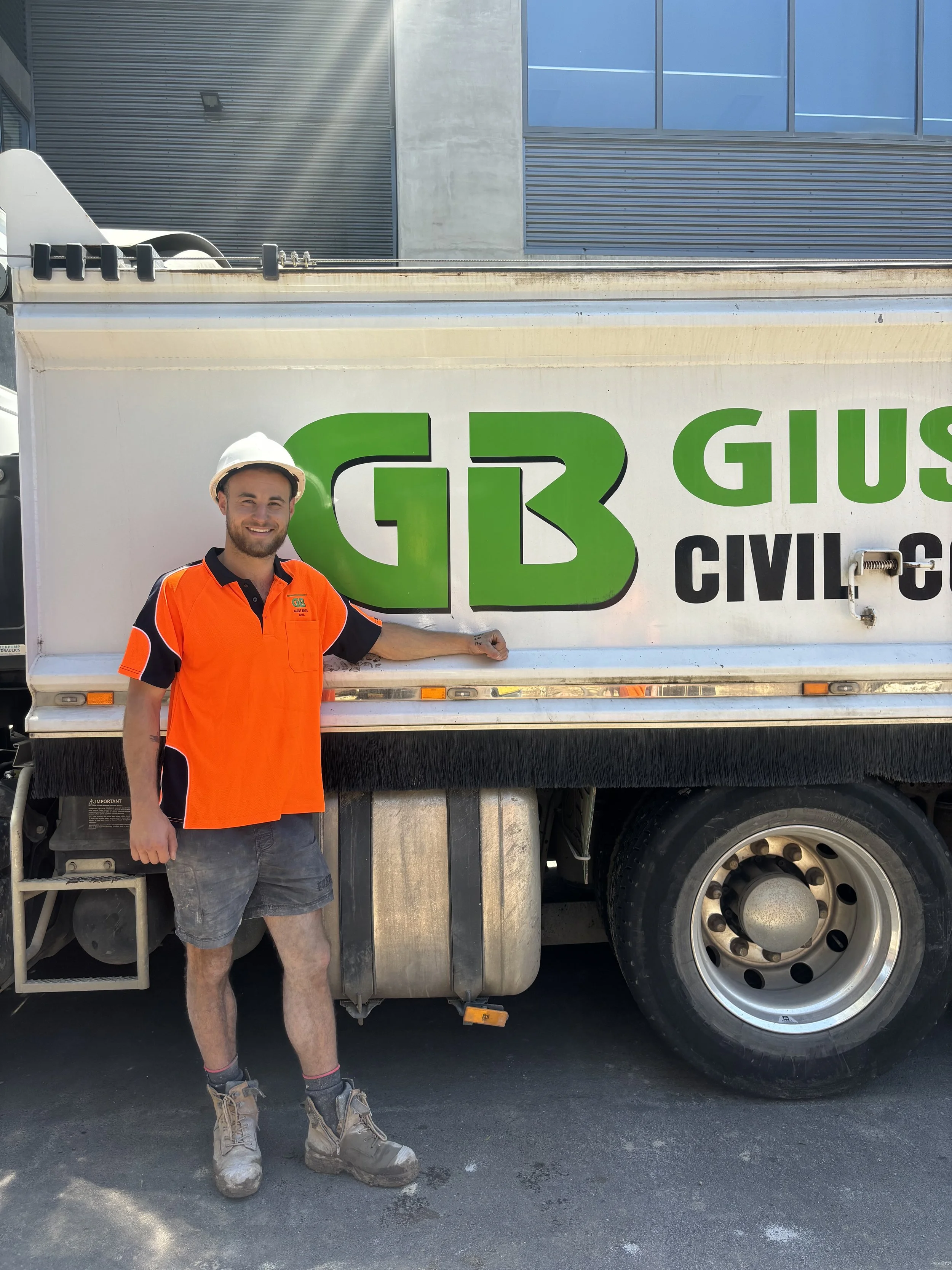 A man wearing an orange safety shirt and a white helmet standing next to a truck with green and black text that reads 'G3 GIUSEPPE CIVIL' on it.
