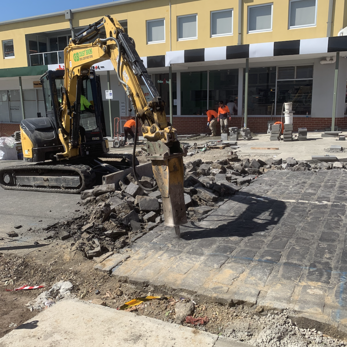 Construction workers and a small excavator removing pavement in front of a commercial building with a yellow facade.