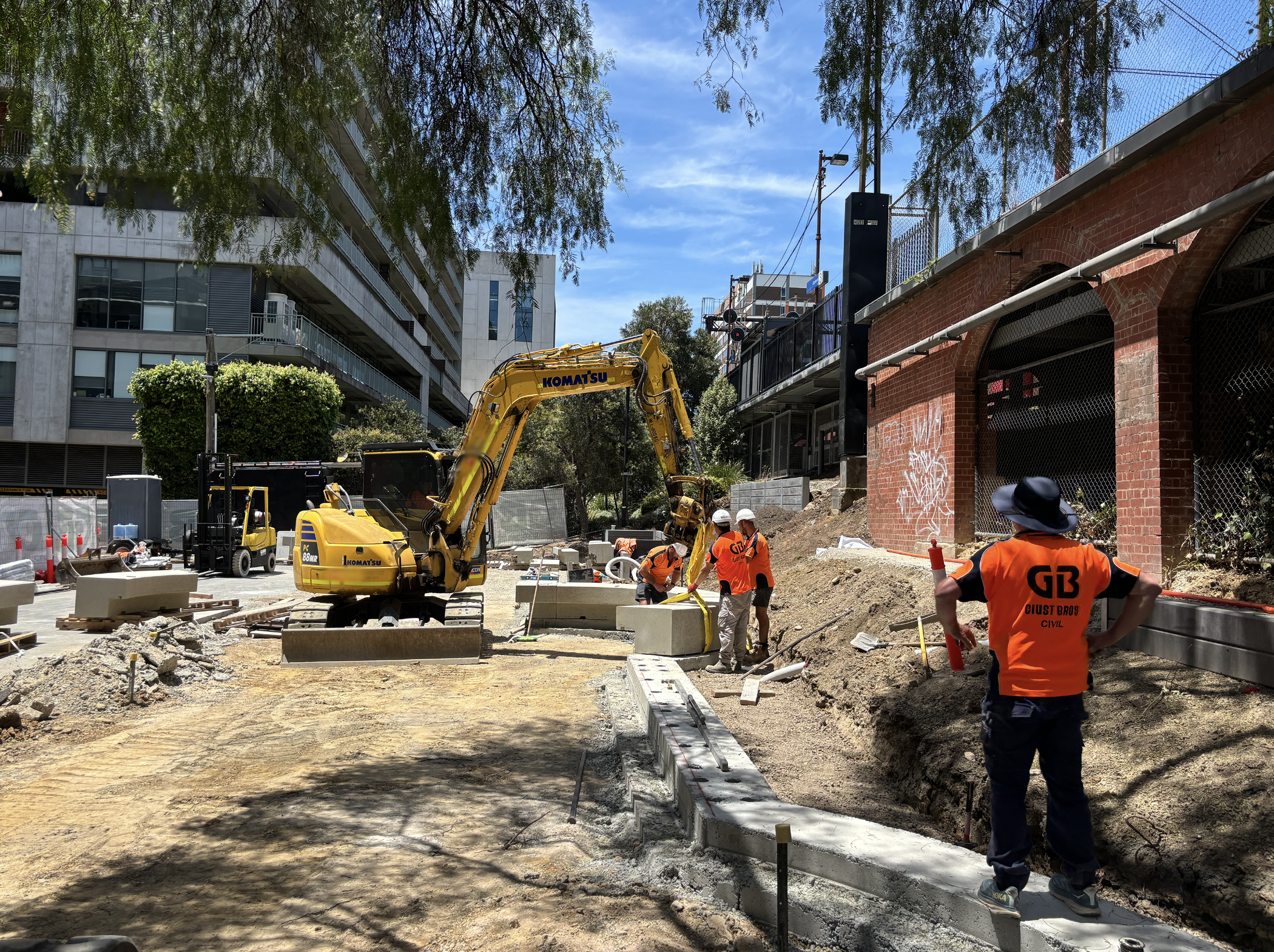 Construction workers in orange shirts and white helmets working on sidewalk project with a yellow excavator and concrete barriers, urban setting with trees and buildings.